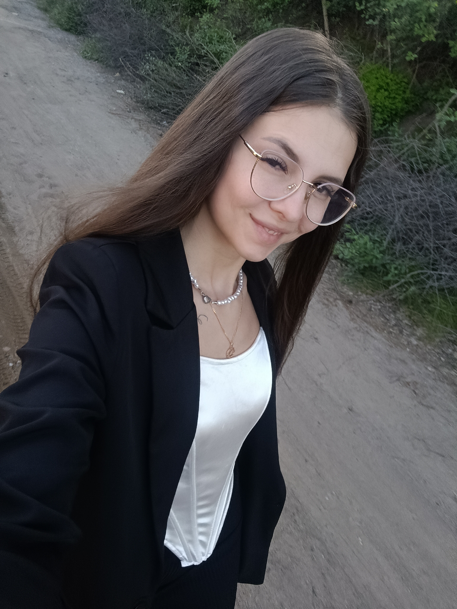 A young woman with long, brown hair and glasses is posing for a photograph on a dirt road. She is wearing a white top, a black blazer, and a pearl necklace with a gold pendant. The road is surrounded by trees and bushes. The location is Unknown, Unknown.