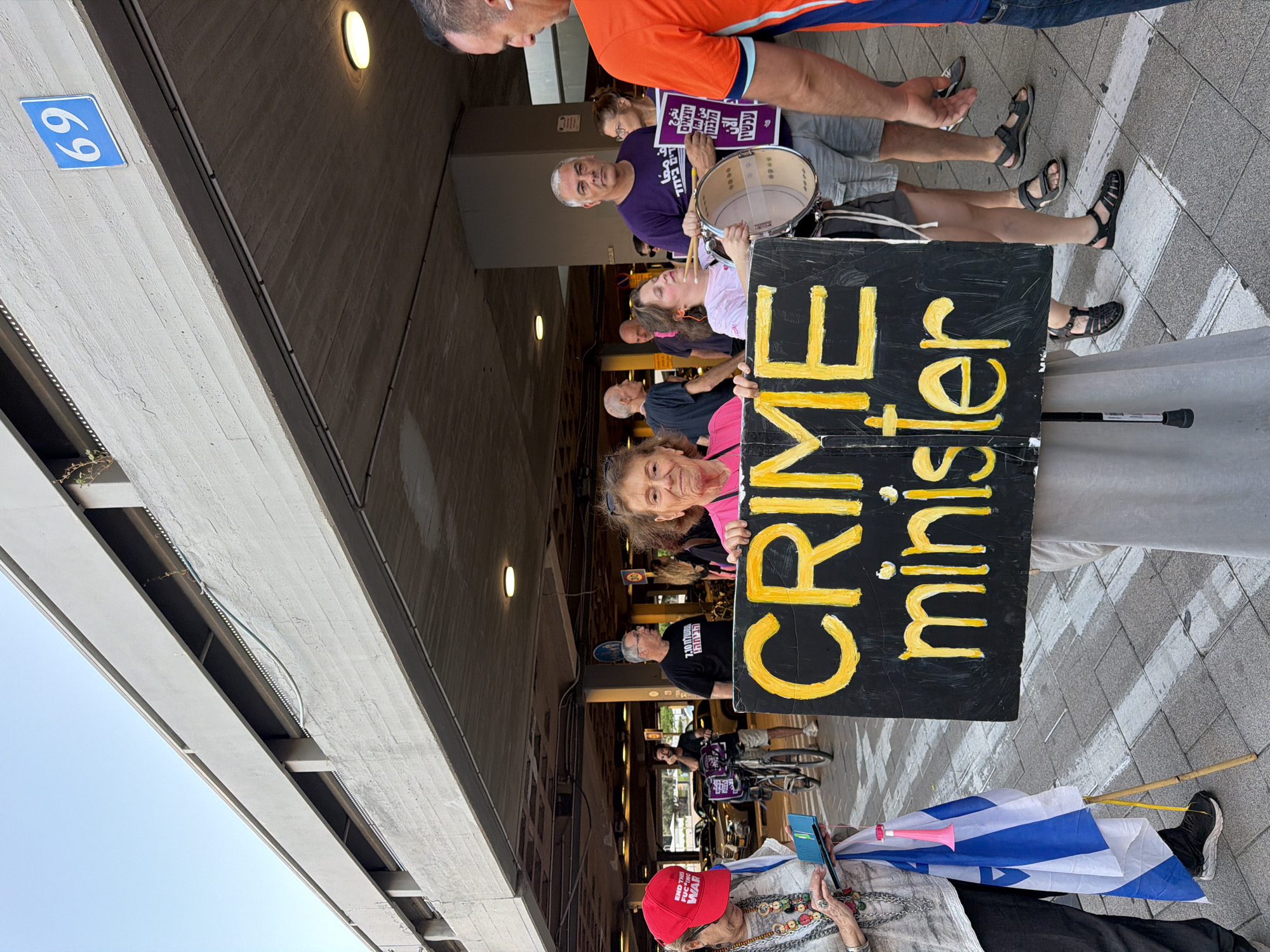 The image depicts an outdoor scene with several people. In the foreground, a woman is holding a black sign with yellow lettering that reads "CRIME minister". To her left, an Israeli flag on a pole is visible. Behind her, a woman is playing a drum with drumsticks. Further back, other individuals are standing, and some are holding signs. One sign is purple with Hebrew text. A man in an orange shirt is partially visible on the far right. The environment appears to be a street or walkway under a concrete overpass, possibly part of a parking garage, with a blue sign displaying the number "69" visible on the upper structure. Cars are parked in the background. The presence of the Israeli flag and the Hebrew text on signs suggests the location is in Israel. While the image doesn't explicitly state Tel Aviv, such overpasses and street layouts are common in urban areas like Tel Aviv.