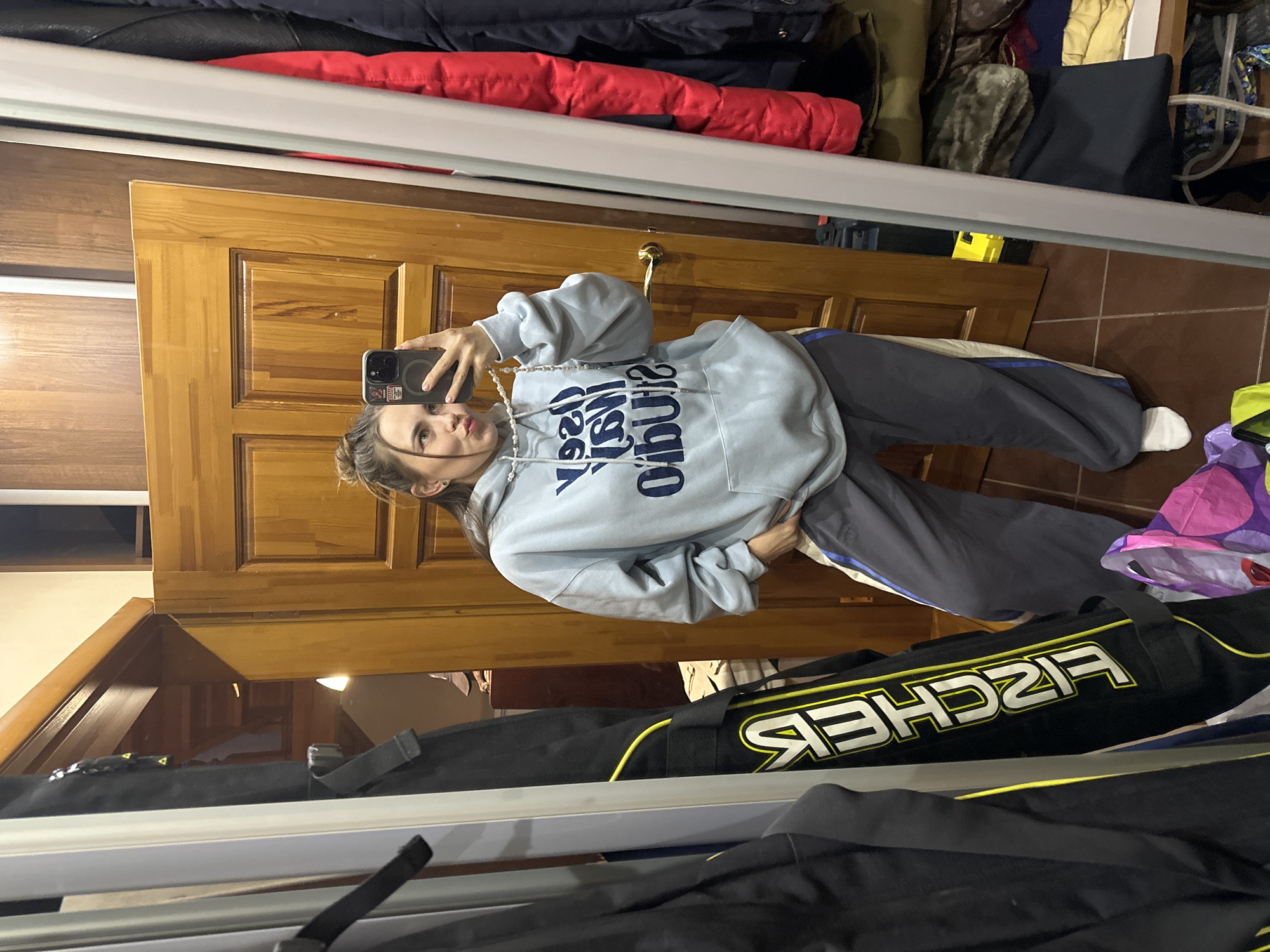 A young woman takes a mirror selfie. She is wearing a light blue hoodie with dark blue writing, dark gray sweatpants with a light stripe, and white socks. She is holding a black smartphone with a dark case. A partially visible doorway is behind her, and various clothing items are stored on a shelf overhead. A black Fischer ski bag is in the foreground. The scene appears to be inside a cluttered room. The location is potentially Ilyinsky, Russia, but this cannot be definitively determined from the image.