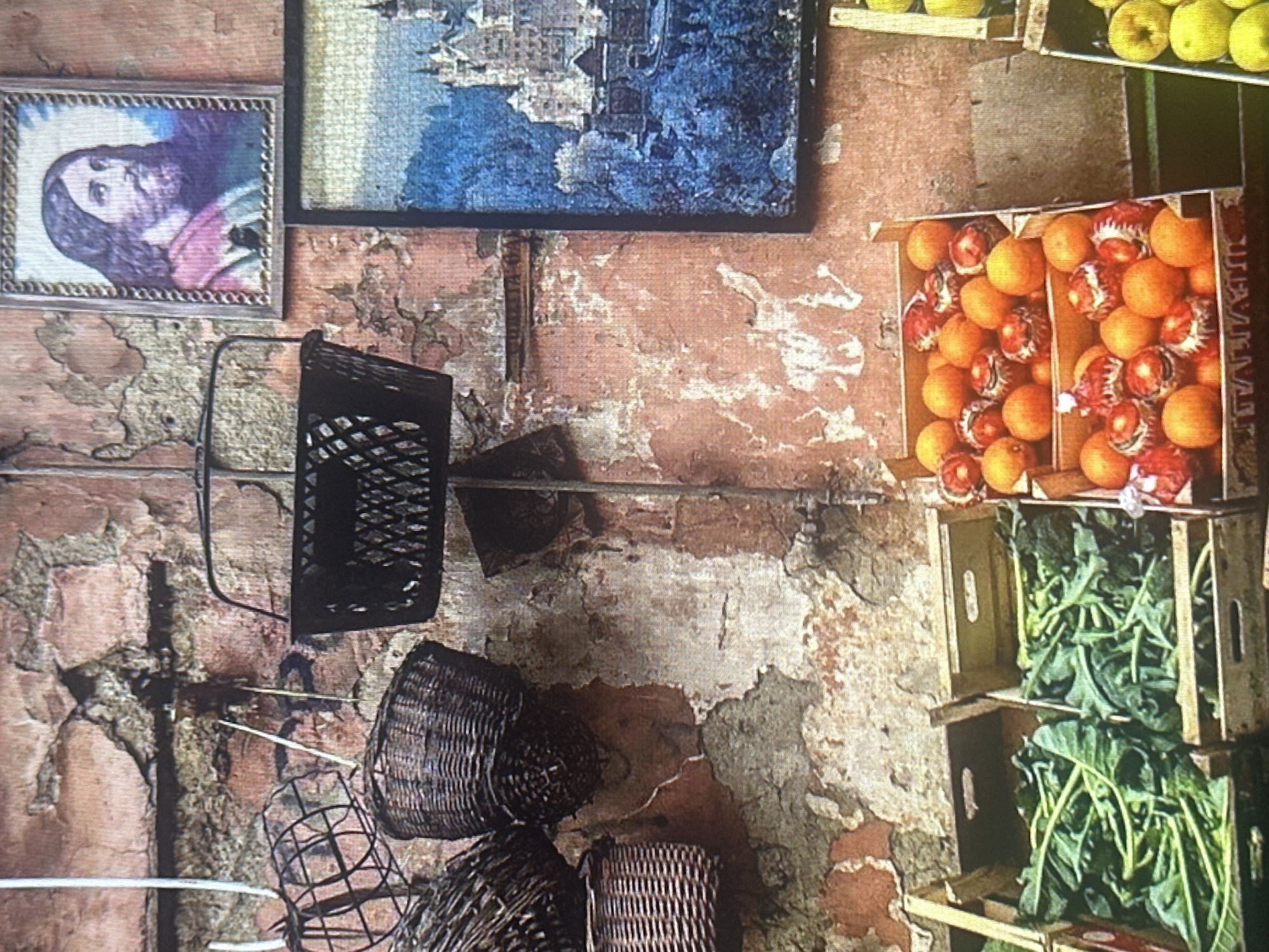 A close-up, eye-level shot captures a rustic outdoor scene, likely a market stall. The background is a textured, distressed wall with peeling pink and ochre paint, revealing patches of grey plaster. Hanging from the wall are several wicker baskets of varying sizes and styles, some appearing weathered. To the right, two framed pictures are mounted on the wall. The upper frame contains a portrait of Jesus Christ. Below it, a larger framed image depicts a picturesque castle set against a vibrant blue landscape. In the foreground, wooden crates are filled with fresh produce. Two crates on the left hold large bundles of leafy green vegetables, possibly broccoli rabe. A third crate, to the right, is overflowing with round, orange fruits, some appearing to be oranges, interspersed with decorative red and white objects. Another crate to the far right is filled with yellow fruits, likely apples. The lighting suggests it is daytime.