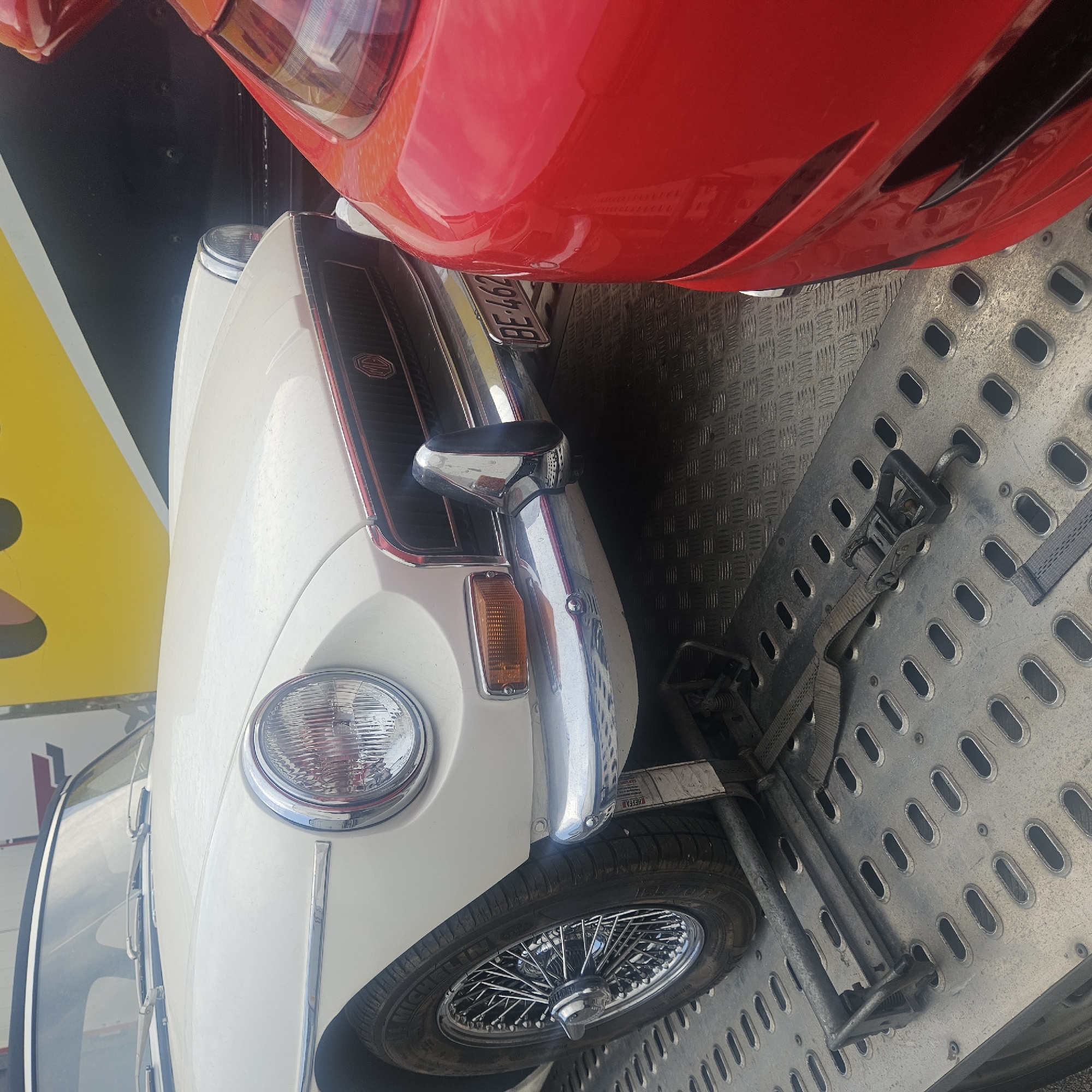 The image shows the front right side of a white classic convertible sports car, possibly an MG MGB, being transported on a flatbed trailer. The car has a chrome bumper, a distinctive headlight with a chrome bezel, and a wire-spoke wheel with a Michelin tire. A license plate that reads "BE-462" is visible on the front of the car. A red sports car is parked to the right of the white car, with its front section visible. The trailer's surface is made of a metal grating with oval-shaped holes, and straps are used to secure the vehicles. In the background, a large yellow and black object, possibly a truck or billboard, is partially visible. The scene is likely in La Jonquera, Spain, a town known for its border crossings and busy commercial activity. No people are visible in the image.