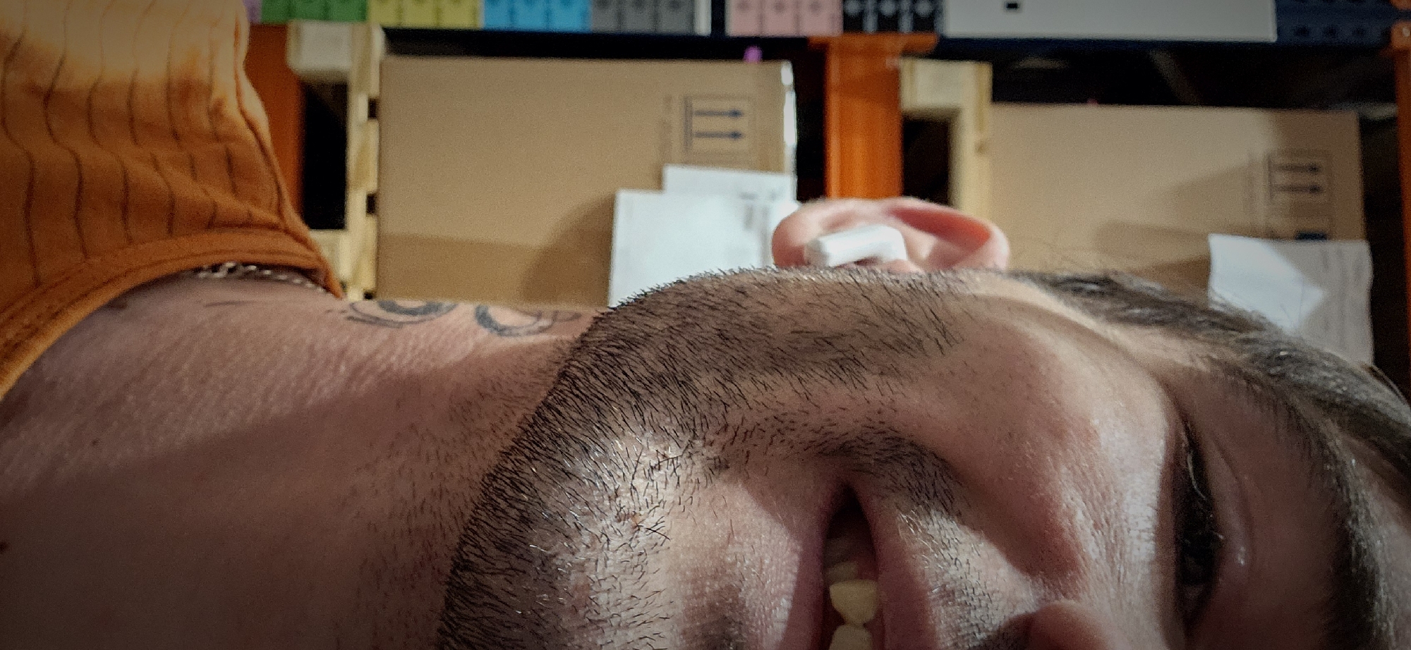 A close-up shot focuses on a man lying down. His head is tilted, revealing his face with partially closed eyes, a visible beard, and a glimpse of his teeth. An object, possibly an earbud, is placed in his ear. He wears an orange top with vertical stripes, and a silver chain is visible around his neck. A tattoo is visible on his upper chest/neck area.
The background is filled with cardboard boxes and shelves. Above the boxes, a shelf with items of varying colors and shapes is visible.
Without additional information, the location in Plovdiv, Bulgaria, cannot be determined.