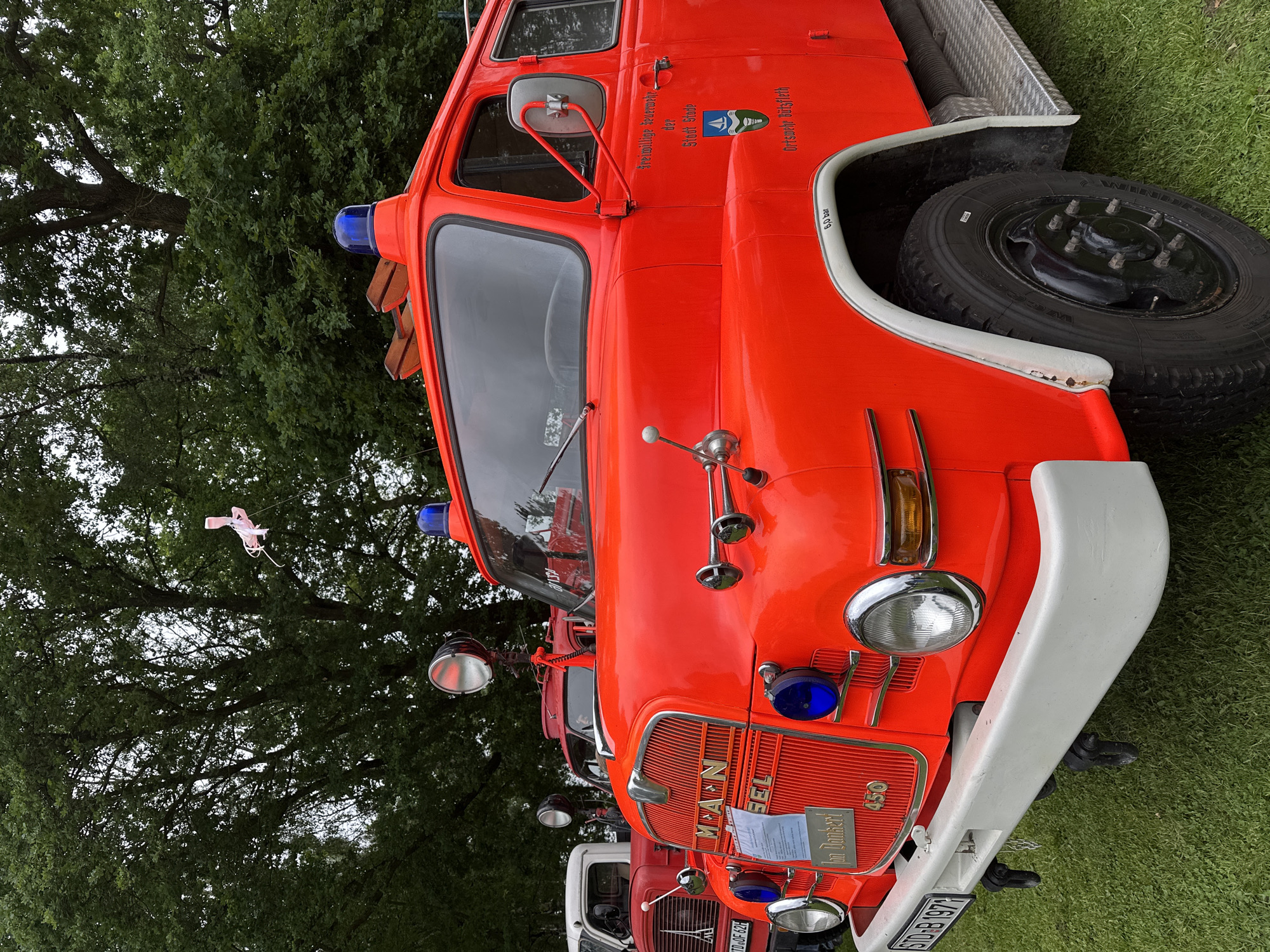The image captures a static display of multiple vintage fire trucks in an outdoor, grassy setting, likely in Deinste, Germany. In the foreground, a prominent bright red MAN fire truck, model 450, is positioned at a front-quarter angle, facing slightly left. The truck features a chrome grille with "MAN" and "SEL" lettering, a white front bumper, and a license plate reading "STD-B 1971". Two round headlights with chrome bezels are visible, flanked by two smaller blue-tinted auxiliary lights below them. Above the driver's side of the cab, a large chrome spotlight is mounted, and two blue rotating emergency beacons are present on the cab roof. Chrome air horns extend from the passenger side fender.
The driver's side door displays "Freiwillige Feuerwehr der Stadt Stade" (Volunteer Fire Department of the City of Stade) and "Ortschaft Bützfleth" (Village of Bützfleth) with a crest depicting a sailboat and green land features. A "6.0 bar" marking is visible near the passenger side wheel well, which has a white trim. The truck's wheels are black with visible lug nuts, and a diamond-plate running board is present on the passenger side.
In the mid-ground, partially obscured and out of focus, are at least two other vintage fire trucks, one red and one white, confirming a collection or event display. One of these trucks has a partially visible license plate "STD-OF 62H". The background is dominated by a dense canopy of large green trees, suggesting a park or rural area, under an overcast sky. A small, light pink, kite-like object with streamers is caught in the branches of one of the trees. The trucks are parked on a well-maintained green grass lawn.