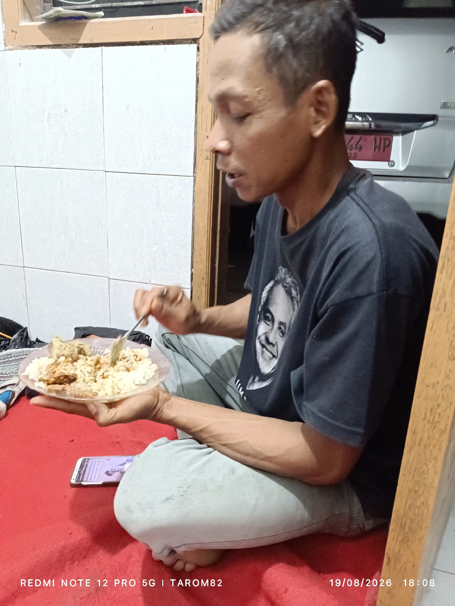 A man is sitting on a red carpet, eating from a clear plastic plate of rice and food. He is wearing a dark t-shirt with a portrait of a smiling man on it and light-colored pants. His right hand is holding a fork and is in the process of taking food from the plate. A smartphone is lying face up on the red carpet to his left. The background consists of tiled walls and a wooden doorframe. The timestamp on the image indicates it was taken on August 19, 2026, at 18:08 in Tarom82, Sidodadi, Indonesia.