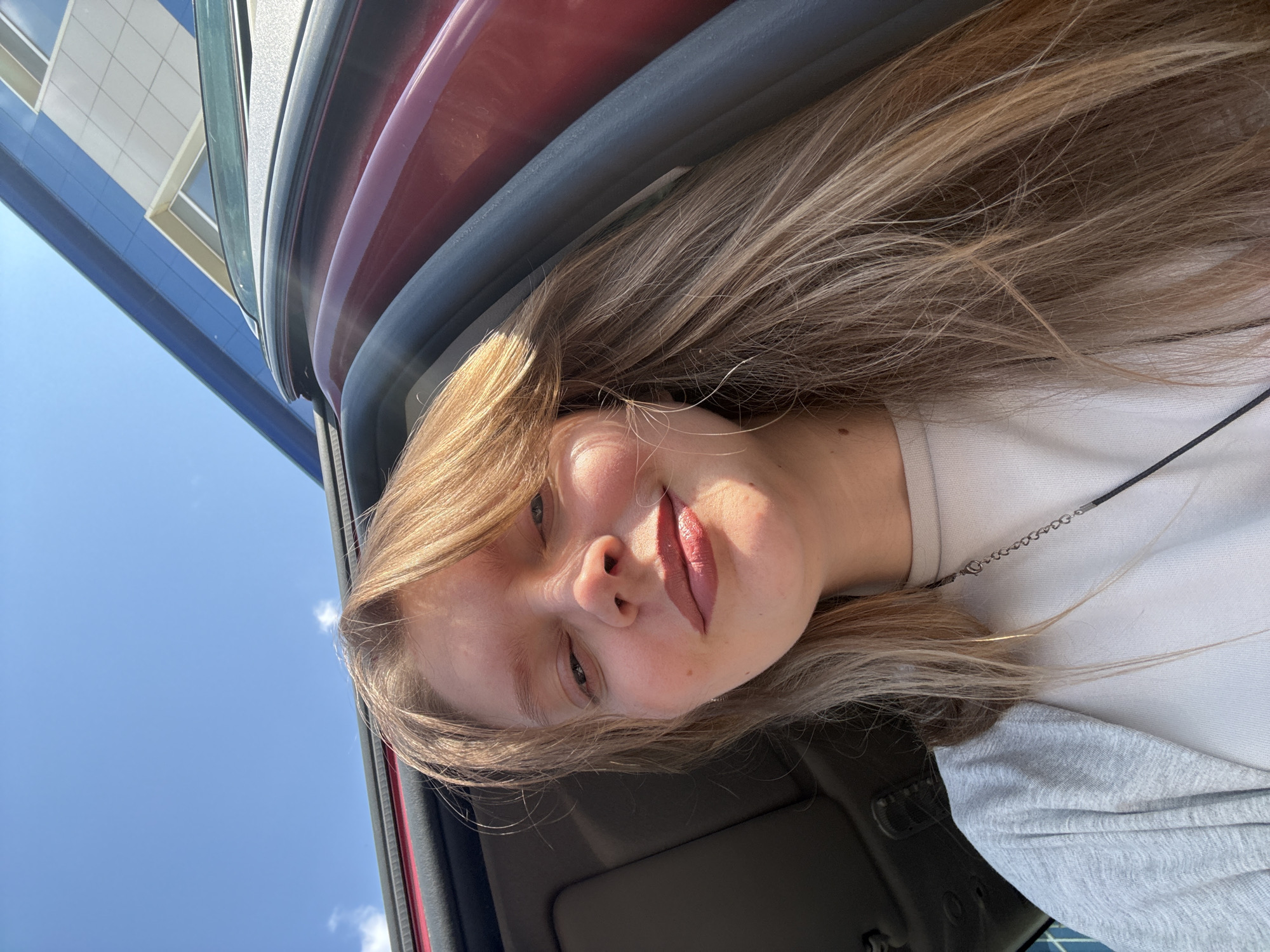 A young woman is taking a selfie from inside a car. She has long, blonde hair and is wearing a white shirt with a gray long-sleeved shirt underneath. She appears to be in the driver's seat. The car door is visible, with the sky and a building visible through the window. The building appears to have blue and white exterior. The image suggests it is a sunny day. The exact location in Izhevsk, Russia, is not discernible from the image alone.