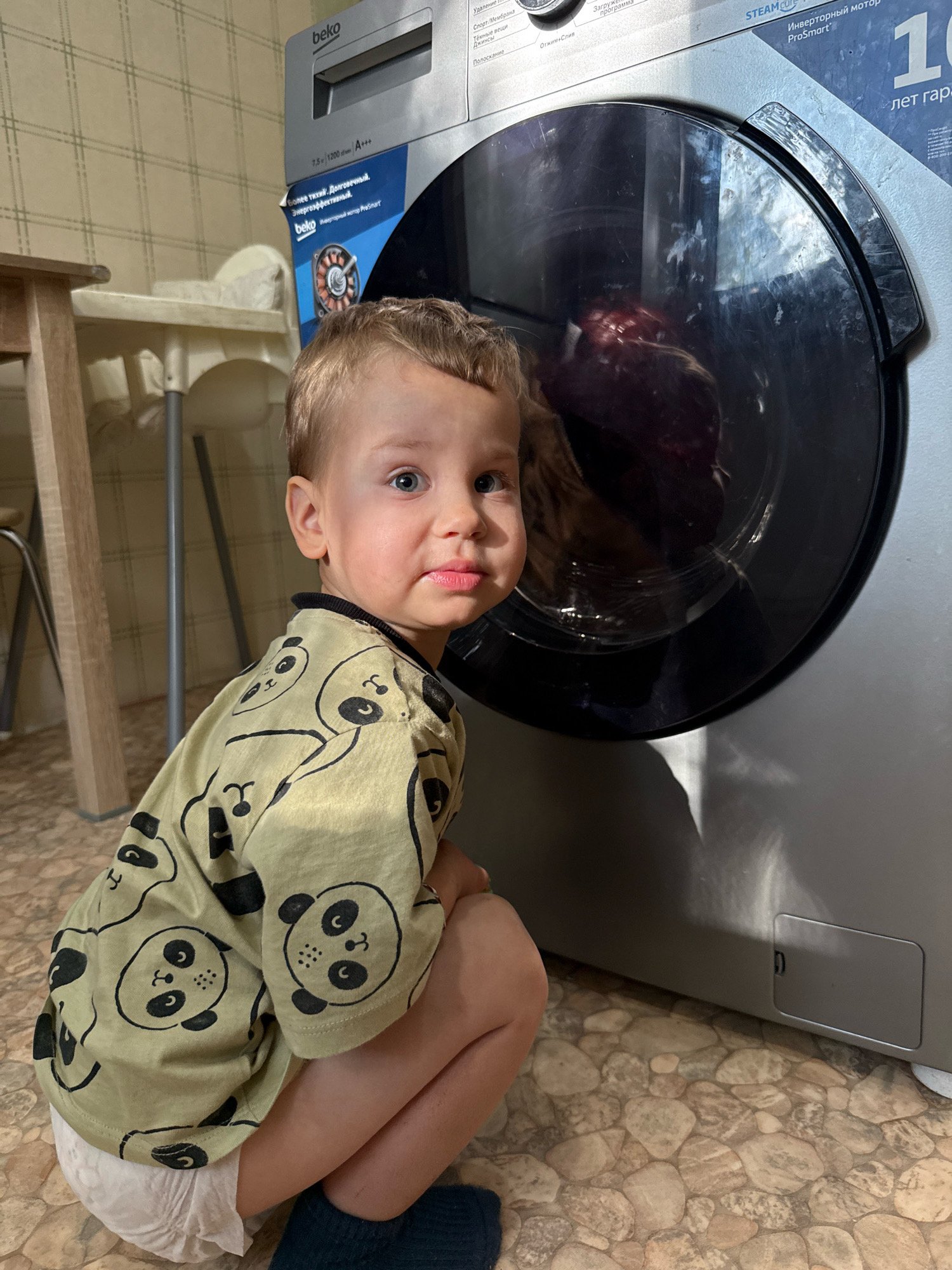 In Rusanovo, Russia, a young child with blond hair and blue eyes kneels on a stone-patterned floor in front of a washing machine. The child is wearing a light green t-shirt with a panda print and a white diaper. The washing machine is silver and has a large, round glass door through which dark laundry is visible. The scene appears to be indoors, with a checkered wallpaper visible in the background and a wooden table and chair to the left. Sunlight streams in, casting shadows on the floor and washing machine.