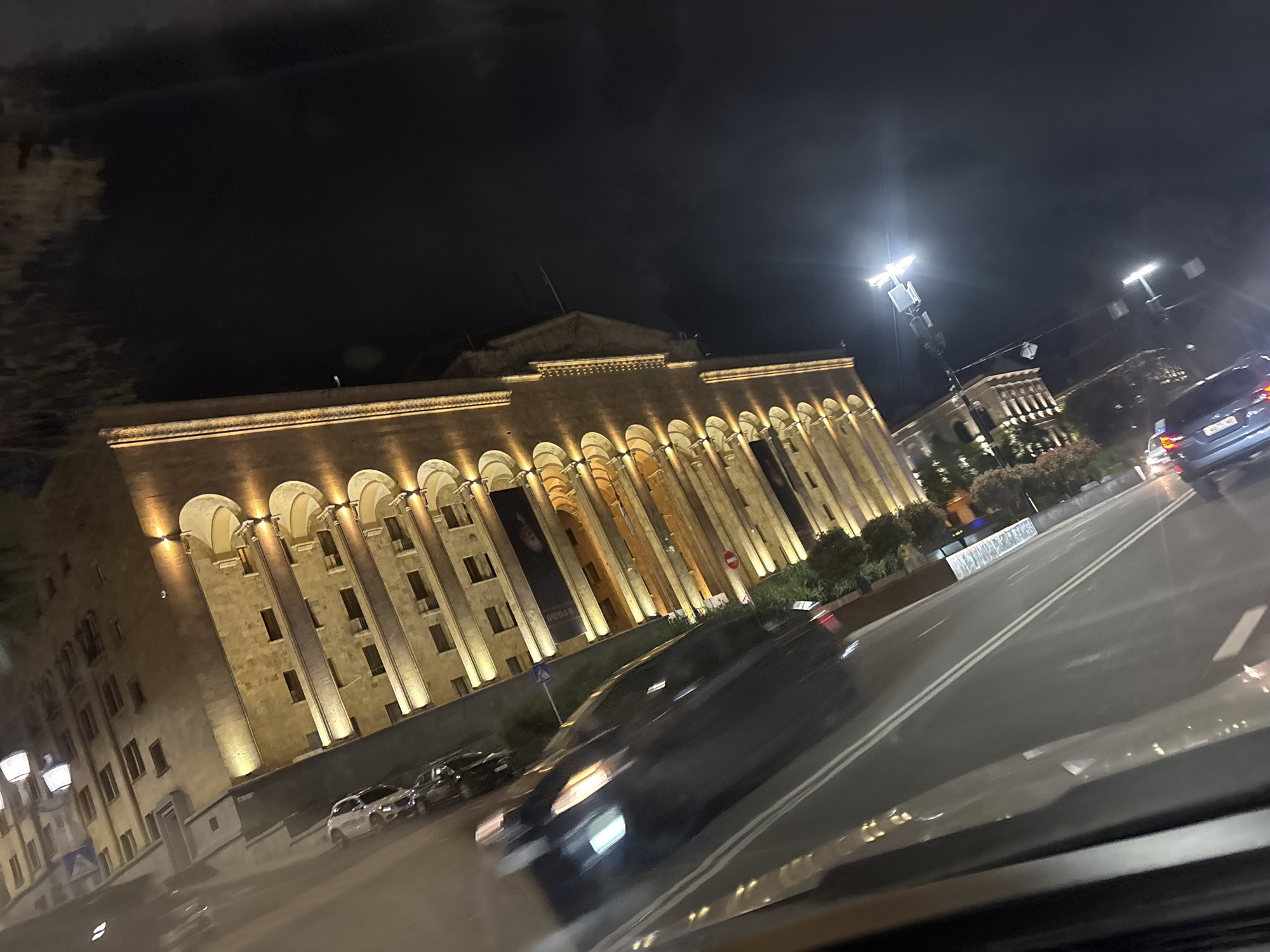 The image presents a nighttime street scene in Tbilisi, Georgia, captured from a moving vehicle. The primary subject is a large, imposing building with classical or neoclassical architecture, characterized by a long facade featuring numerous arched openings and prominent pilasters or columns. This building, resembling the Parliament of Georgia, is illuminated with warm uplighting, making it stand out against the dark night sky. A large banner depicting a person's face and displaying Georgian text, possibly related to an exhibition or historical context (with "ისტორია" - history - potentially visible), hangs on the building's facade.
In the foreground and mid-ground, a multi-lane asphalt road is visible, clearly marked with white double lines. Several vehicles populate the scene; some cars are in motion and appear blurred, while others are parked along the street on the left side of the frame. Bright streetlights line the road, casting strong illumination and creating lens flares.
Additional details include trees and bushes situated along the roadside and in front of other buildings in the background. A blue road sign with a white arrow is visible on the left, and a small, indistinct red and white circular sign is near the main building. The dark sky confirms the nighttime setting. The composition is slightly tilted, consistent with a photograph taken from a moving vehicle, with reflections visible on what appears to be a car's dashboard or windshield in the lower part of the frame. The scene conveys a sense of a well-lit, active urban environment after dark.