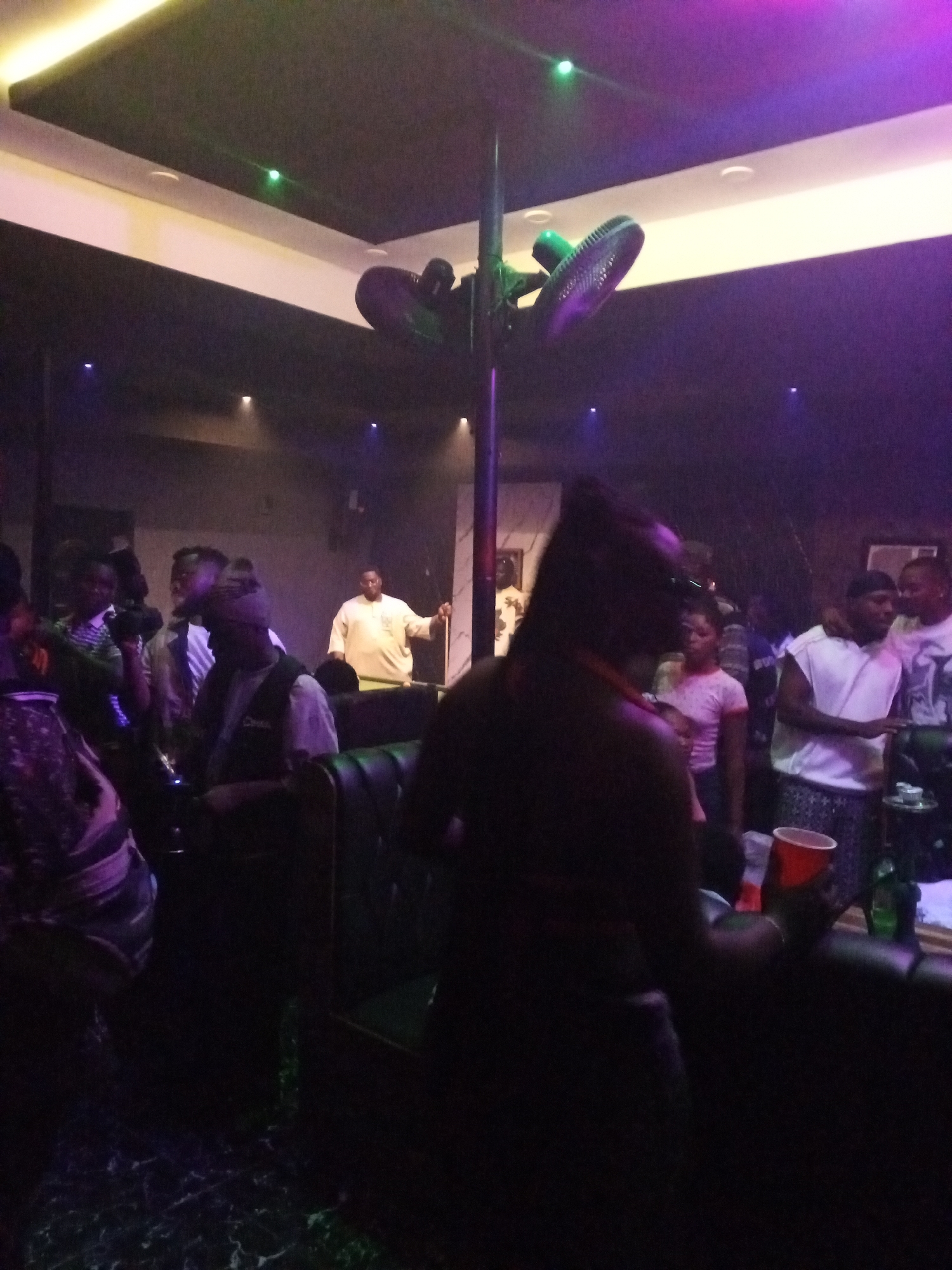 The scene depicts a dimly lit indoor space, likely a nightclub or lounge in Lokoja, Nigeria. A central pole is visible with a person positioned near the top, appearing to hang or climb. Several individuals are gathered around tables and standing, some holding cups or drinks. A pool table is partially visible in the background. The room features a decorated ceiling with recessed lighting and various decorative elements. The environment appears crowded and social, with a mix of actions and interactions amongst the people present.