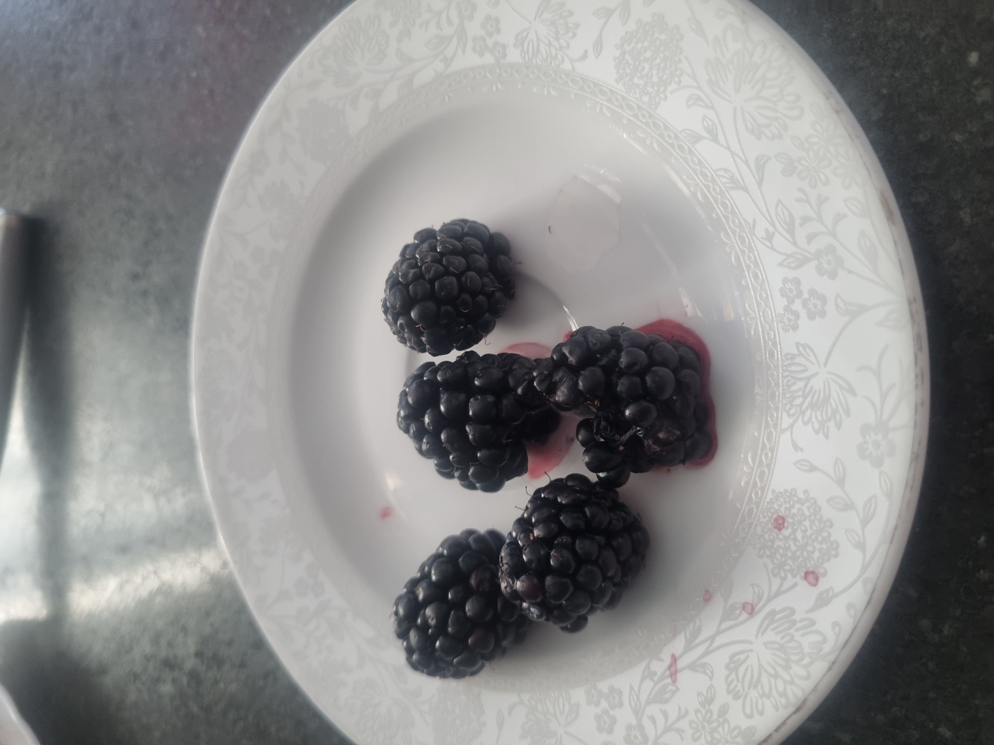 Five blackberries are arranged on a white plate with a floral pattern. The plate is on a dark, speckled surface, likely a countertop or table. There are some red stains on the plate, suggesting the blackberries have released juice. The lighting is overhead, casting faint shadows. The image quality is decent, showing the texture of the berries and the detail of the plate's design. There are no people or indications of the city of Samara, Russia, in the image.