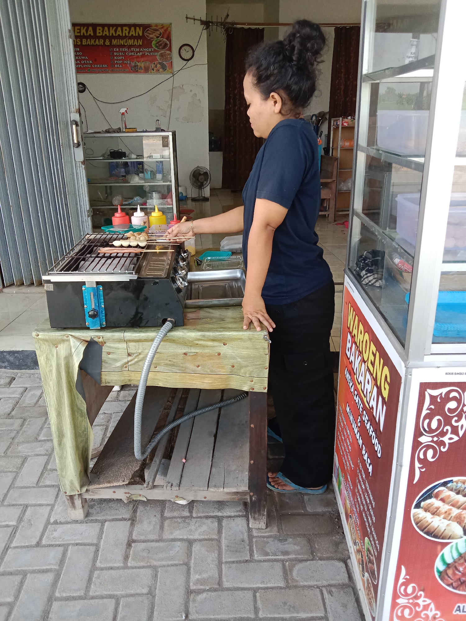 A woman is grilling meat on a portable grill in Turunsih, Indonesia. She is standing to the right of the grill, with her right hand holding a skewer of meat. The grill is on a wooden table covered with a patterned cloth. Behind the grill, there is a glass display case with various food items. To the left of the grill, there is a corrugated metal gate. The ground is paved with interlocking bricks. A sign above the grill advertises "Aneka Bakaran & Minuman" which translates to "Various Grilled Foods & Drinks". Another sign on the right side of the image reads "WAROENG BAKARAN", indicating a food stall. The scene appears to be outdoors, possibly in front of a shop or in a market area.