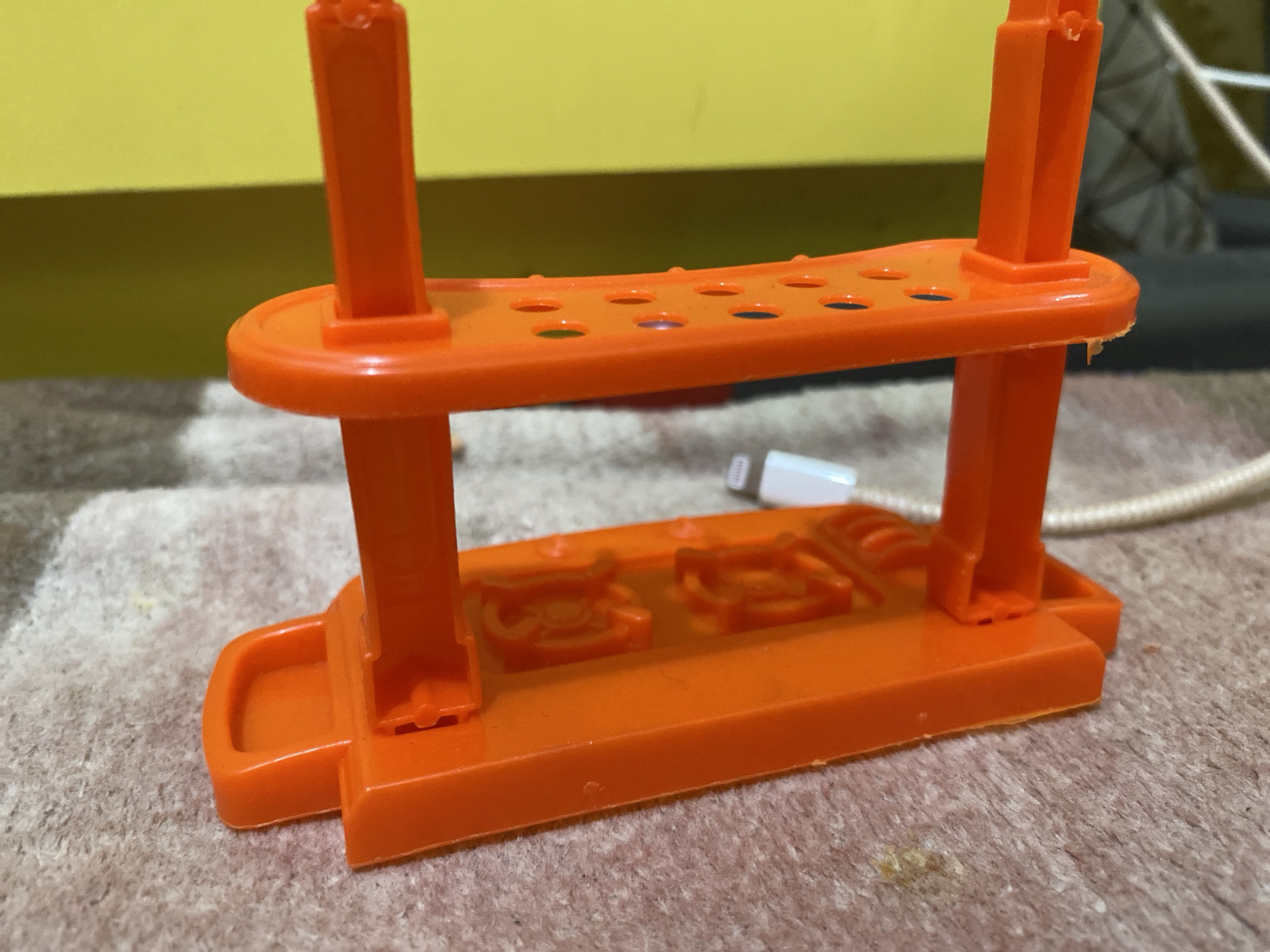 The image shows an orange plastic stand for holding small items, possibly test tubes or similar cylindrical objects. The stand has a base with indentations, two vertical supports, and a top platform with multiple circular holes. A white cable is visible behind the stand. The stand is placed on a textured surface that appears to be a carpet or mat. In the background, a yellow wall is visible. The scene is located in Lambaro Kueh, Indonesia.