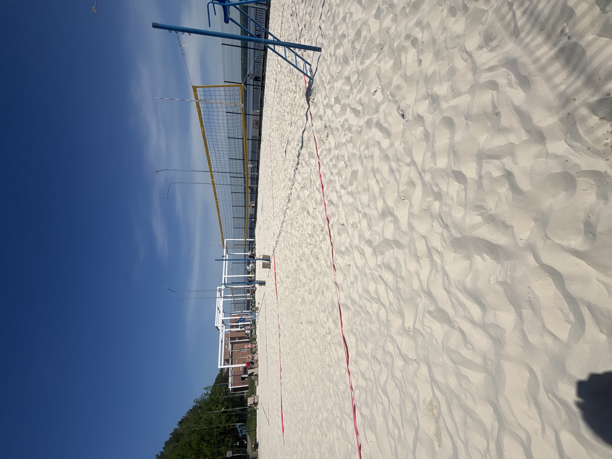 The image shows a beach volleyball court located in the city of Chaikovsky, Russia. The court is situated on a sandy beach, bordered by trees and buildings. A volleyball net is present, dividing the court into two halves. The lines of the court are visible. The sky is blue, and there is sunlight present. Some people are located near the court.