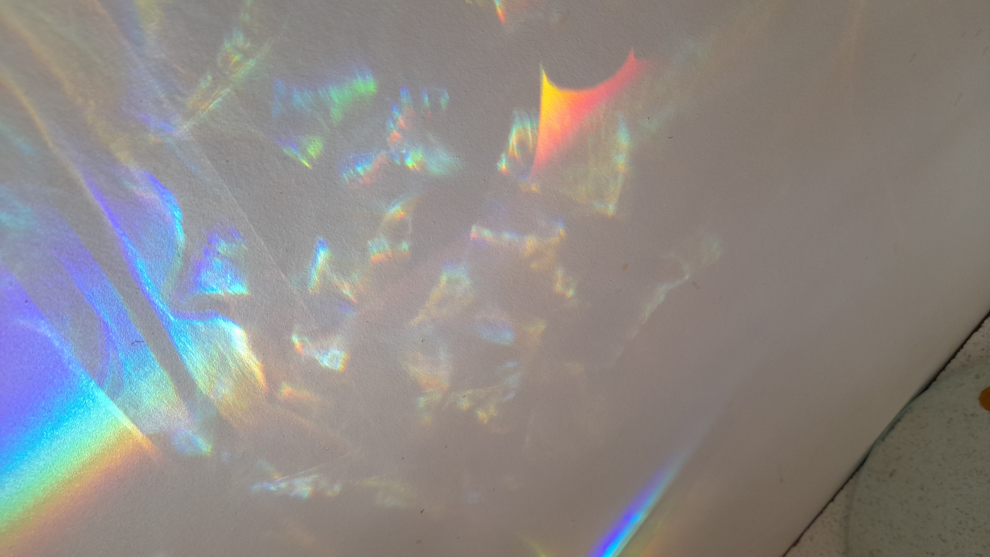 The image shows refracted light patterns on a white or light-colored wall. The light appears to be from a prism or reflective surface, casting rainbow-like colors and shapes across the surface. The image is taken at an oblique angle, and the corner of the room is shown in the bottom right corner. The scene is simple, dominated by the interplay of light and color on the plain wall. It does not appear that the image was taken in Saint Petersburg, Russia.