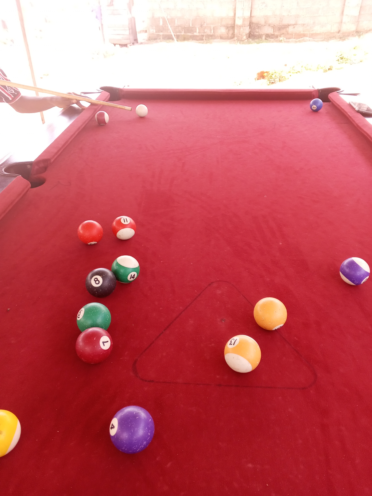 This image depicts a red billiard table in Lokoja, Nigeria, likely outdoors given the bright, blown-out background suggestive of harsh sunlight and a rough, possibly brick, wall. The table is set up for a game with several billiard balls scattered across its surface. In the foreground left, a portion of a person's arm and hand is visible, holding a billiard cue and positioned to strike a white cue ball. Adjacent to the cue ball is a red and white striped billiard ball.
The billiard balls present include: a solid blue ball with the number '2', a solid purple ball with the number '4', a solid yellow ball with the number '13', a solid yellow ball with the number '9', a solid red ball with the number '1', a solid green ball with the number '6', a solid black ball with the number '8', a solid green ball with the number '14', a solid red ball with the number '11', and a solid red ball. Additionally, there's a striped purple and white ball and a solid yellow and white ball. The balls are arranged in a way that suggests a game is in progress or has recently been played, with some balls clustered together and others spread out. A triangular racking outline is faintly visible on the felt. The overall environment appears to be informal, possibly a backyard or a casual outdoor recreational area in Lokoja.
