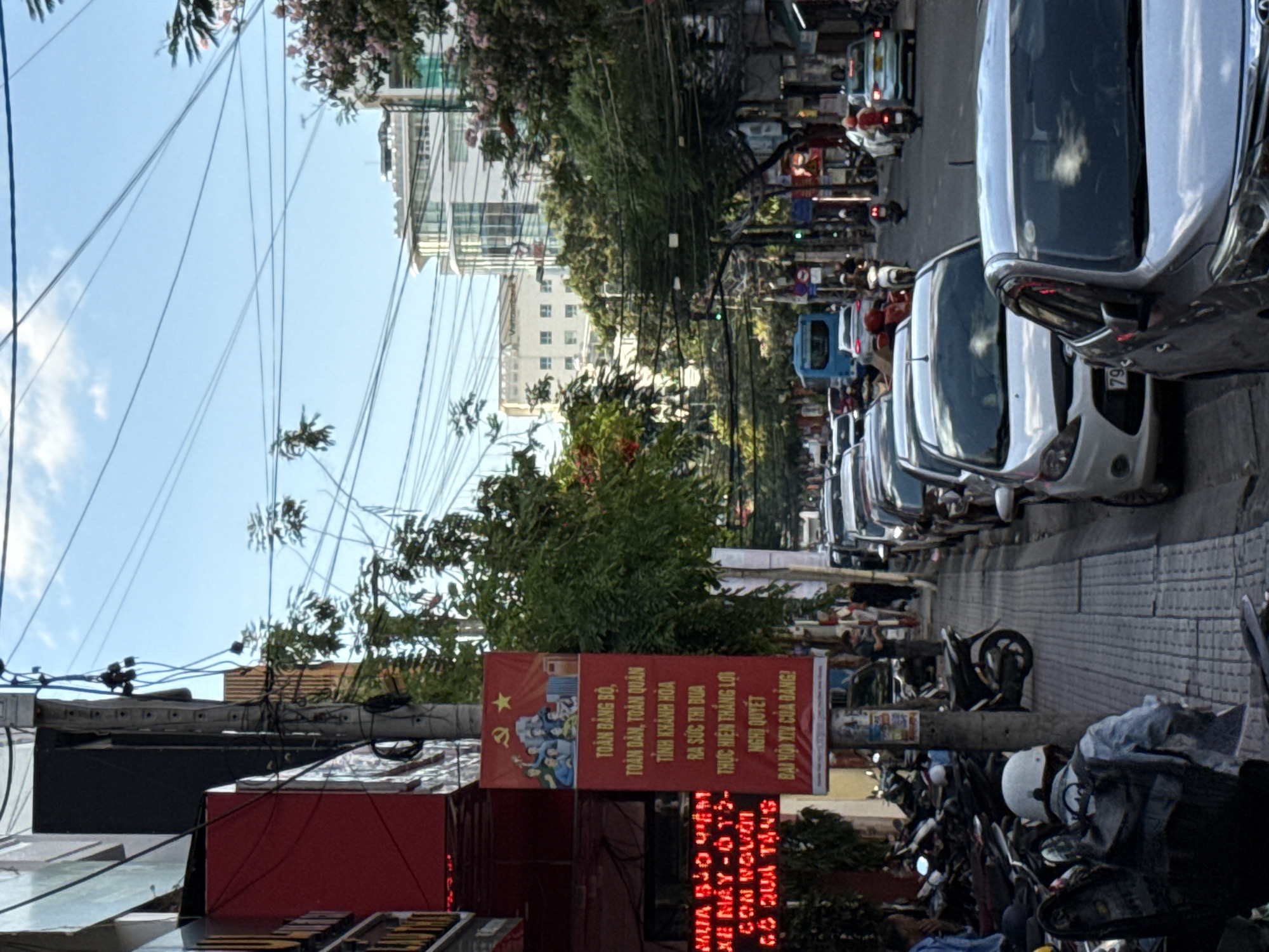The image depicts a street scene in Nha Trang, Vietnam, on a sunny day. A vertical banner with Vietnamese text and communist symbols, including a hammer and sickle with a star, hangs from a utility pole. The banner features an illustration of people holding their hands up in celebration. Below the illustration, Vietnamese text reads: "TOÀN ĐẢNG, TOÀN DÂN, TOÀN QUÂN TỈNH KHÁNH HÒA RA SỨC THI ĐUA THỰC HIỆN THẮNG LỢI NGHỊ QUYẾT ĐẠI HỘI XIV CỦA ĐẢNG!". To the left of the banner, a red sign displays Vietnamese text in yellow, potentially advertising goods or services.
The street is lined with parked motorcycles and several cars. In the foreground, a person wearing a white helmet and a backpack is seated on a motorcycle, facing away from the camera. Further down the street, traffic, including cars and motorcycles, is visible. A blue bus is also present in the flow of traffic. The environment is characterized by buildings, trees with foliage, and a clear blue sky with some clouds. Utility wires are strung across the street.