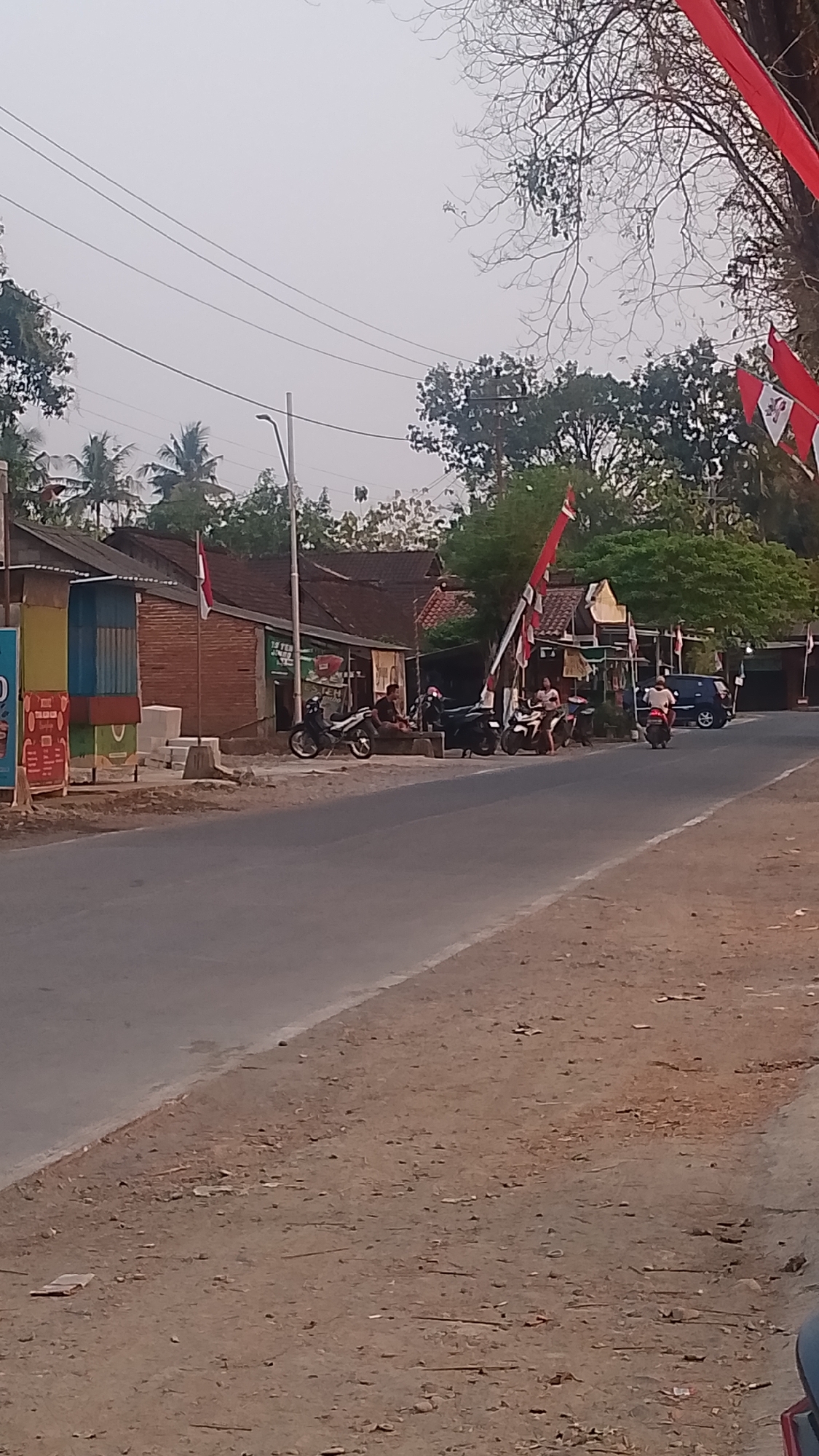 The image depicts a roadside scene in Karanglangu, Indonesia, likely during the day with overcast skies. A paved road runs diagonally from the bottom left to the middle right of the frame. The left side of the road features a dirt shoulder with scattered debris and vegetation. Along the right side of the road, several small buildings with brick and corrugated metal construction are visible. A flagpole stands near one of the buildings, adorned with a red and white Indonesian flag. Red and white banners are also strung across the street.
Several individuals are present, some seated and others on motorcycles. Two motorcycles are parked in front of the buildings, and two more are in motion on the road. One dark blue car is parked further down the road. The environment is characterized by trees, including palm trees, and a general sense of a rural or semi-urban setting. Power lines are visible overhead.