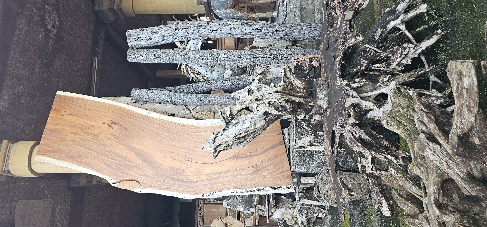 The image depicts a cluttered display of natural and crafted wood elements, likely situated in the Ubud District of Indonesia. The most prominent feature is a large, horizontally-oriented slab of polished brown wood, possibly a tabletop or counter surface. Behind this slab are several bundles of thin, gray-toned, bark-covered branches, appearing to be stacked. Interspersed among these are weathered, tangled pieces of driftwood and root systems, some exhibiting signs of moss or lichen growth. To the right of the frame, darker, more heavily weathered root formations are visible. The background includes hints of stonework and architectural details suggesting an indoor or semi-enclosed space.