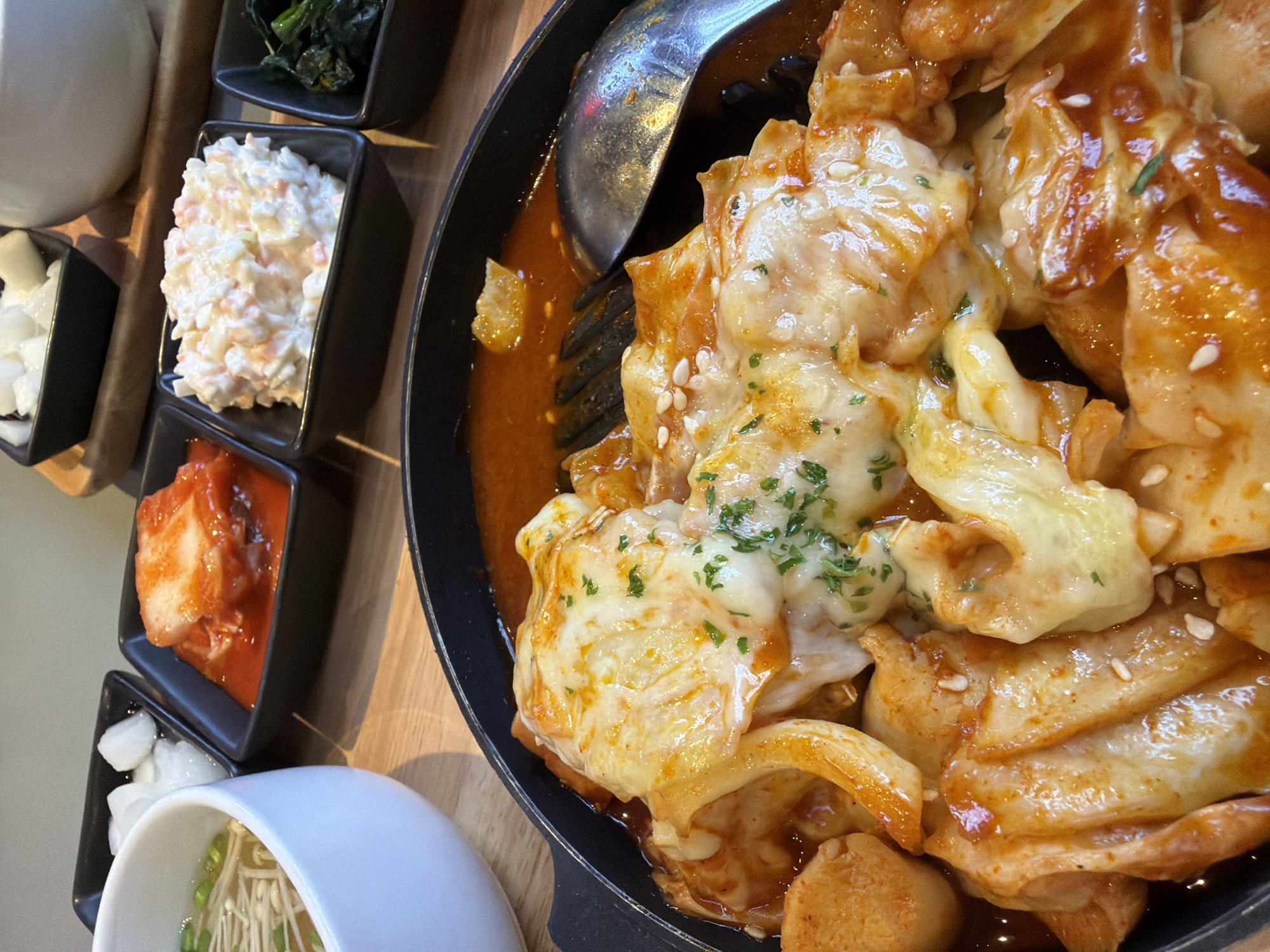 This is a close-up, eye-level shot of a Korean meal served in small black bowls and a large black pan. The pan contains tteokbokki, a Korean rice cake dish, covered in melted cheese and garnished with chopped parsley and sesame seeds. To the left of the pan is a white bowl of soup with enoki mushrooms and green onions. Behind the soup are three small black bowls containing various side dishes: pickled radish, kimchi, and coleslaw. A fourth black bowl, mostly out of frame to the right, appears to contain cooked greens. The dishes are arranged on a wooden table. The background shows a blurred out portion of a restaurant interior. There is no indication of the specific location in Bangkok, Thailand.