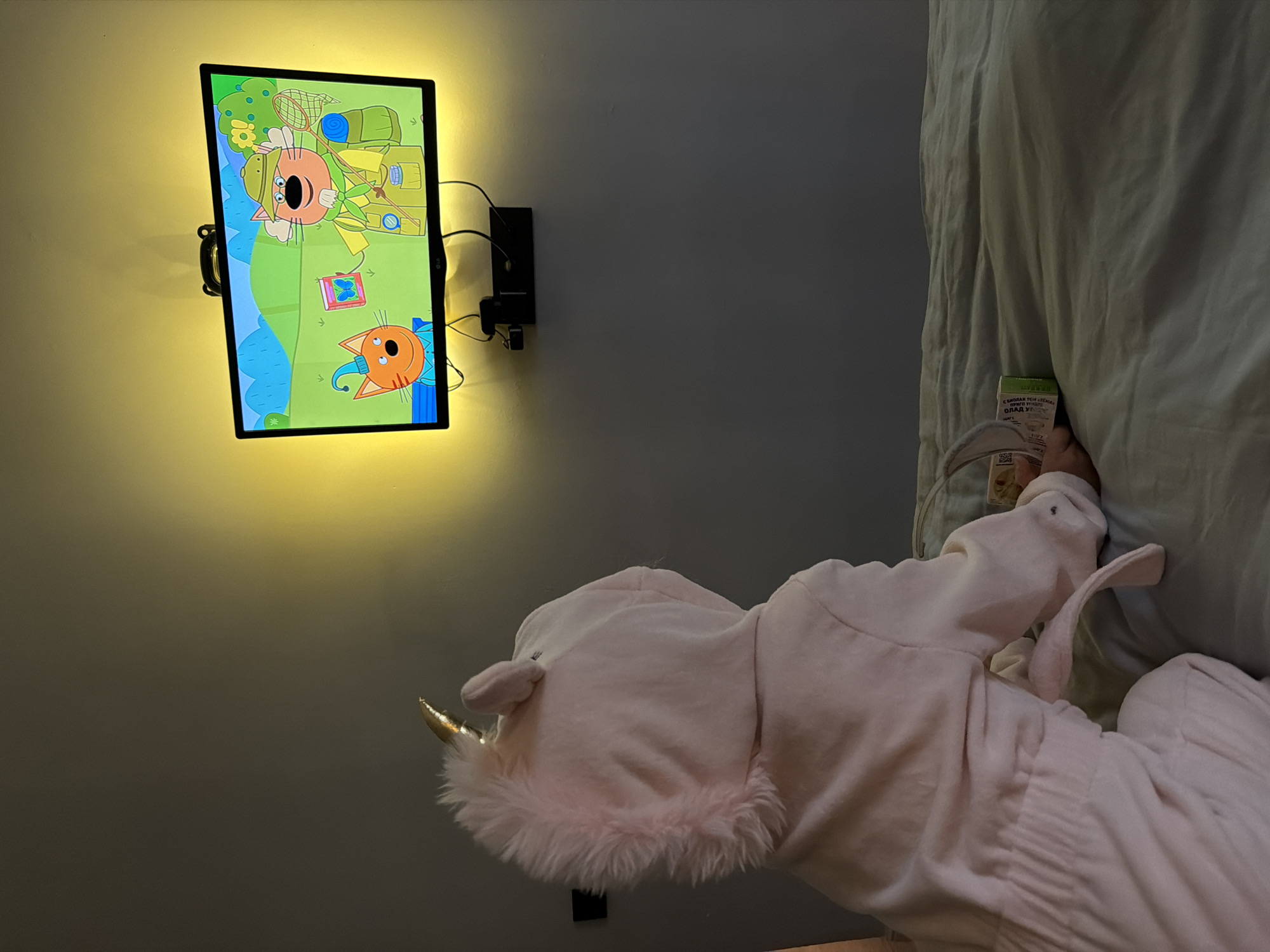 A person wearing a pink unicorn costume sits on a bed, facing a television screen. The television displays an animated scene featuring two anthropomorphic cats. The room is dimly lit, with a warm yellow light emanating from behind the television. A box of snacks is held by the person on the bed. The location is identified as a bedroom in Khimki, Russia.