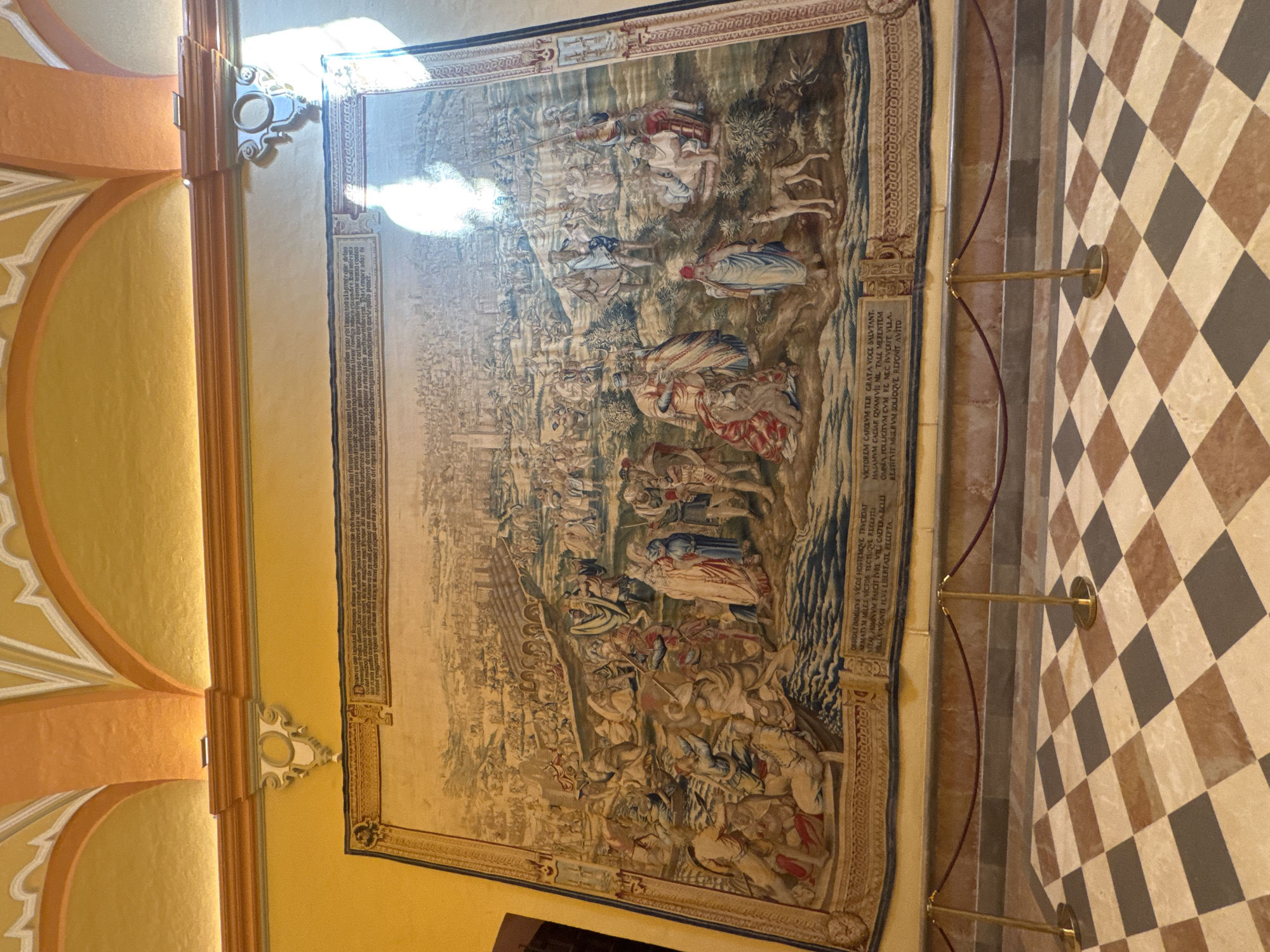 This is a full-shot, eye-level, indoor photograph of a large, historically significant tapestry depicting a battle scene, displayed on a yellow wall in the Alcázar of Seville, Spain. The tapestry, rendered in muted tones of beige, brown, blue, and red, is densely populated with figures engaged in combat. In the foreground, a group of individuals in medieval attire are fighting in a body of water, with some appearing to be on rafts or in shallow boats. Several figures in the middle ground are standing and observing the conflict, some in elaborate robes. In the background, a sprawling city with numerous buildings and structures is visible, suggesting a siege or the aftermath of a battle. The tapestry is bordered by an ornate frame. Below the tapestry, a velvet rope barrier on brass stanchions separates the viewer from the artwork. The floor is a checkerboard pattern of beige, brown, and gray tiles. The ceiling above features orange and yellow arched architectural elements.