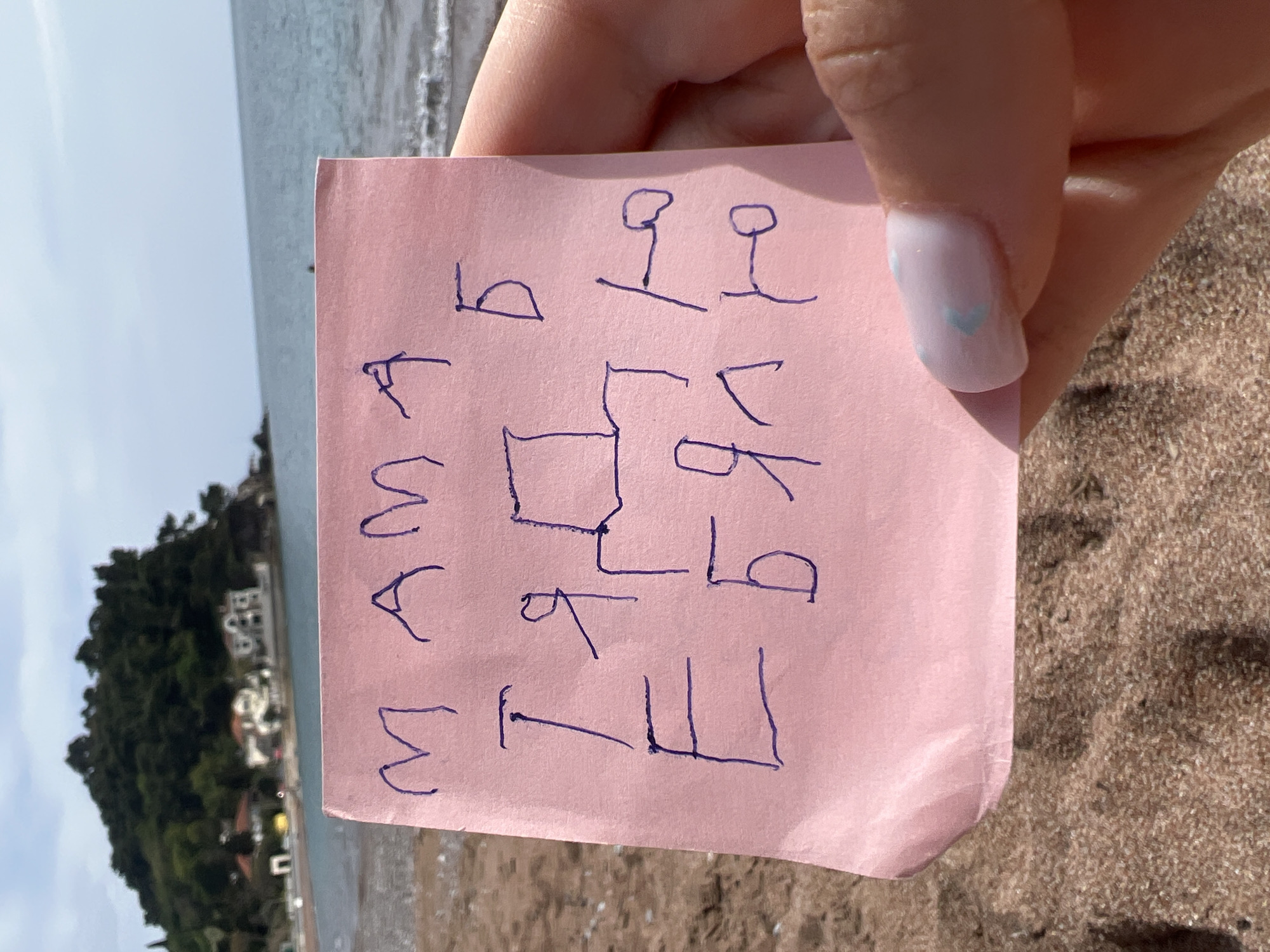 A close-up photograph captures a person's hand holding a small, pastel pink piece of paper adorned with handwritten symbols in blue ink. The scene is set on a sunny beach, with the background revealing the characteristic landscape of Petrovac centar, Montenegro.
The hand, with light pink/white fingernails featuring a subtle blue heart design on the thumbnail, gently grips the top right corner of the slightly creased paper. On the paper, vertically aligned on the left side, are three stylized symbols resembling an inverted 'M' or 'W', with the middle one appearing as an inverted 'V' or Greek Lambda (Λ). To the top right, two identical symbols are visible, each formed by a small loop or circle positioned above a short horizontal line. The central and right sections of the paper contain a cluster of interconnected geometric and curved shapes. These include squares, lines, and forms that vaguely suggest letters such as 'b', 'g', 'p', 't', 'k', and 'E', rendered in a simplistic, blocky, or abstract style, possibly indicating a drawing or a personal code.
The background shows a tranquil beach environment. Golden-brown sand fills the foreground, leading to the calm, light blue waters of the sea. Further in the distance, a verdant hill or mountain rises, dotted with several white buildings, typical of a Mediterranean coastal town. The bright illumination indicates it is daytime with clear weather, suggesting a peaceful and relaxed setting for this activity.