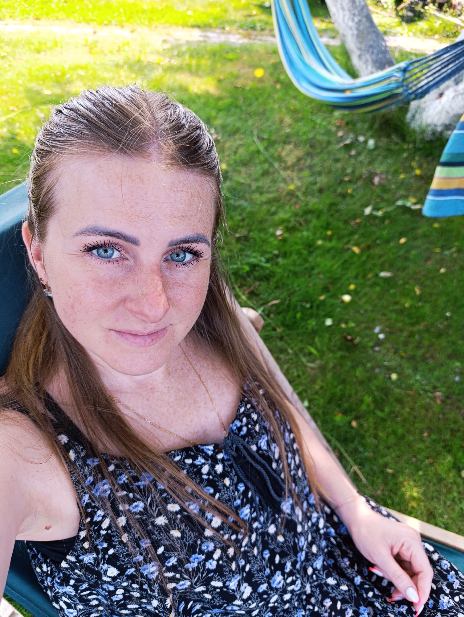 A woman with long brown hair and blue eyes, wearing a black floral dress, sits in a chair outdoors in a grassy area. A striped hammock is visible in the background, suspended between two trees. The overall lighting suggests it is daytime.