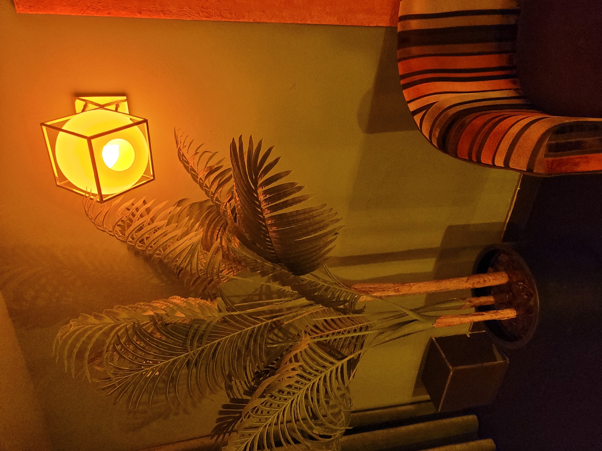 The image depicts an interior scene with a palm plant in a pot, a modern hexagonal wall lamp, and a corner of a striped armchair. The wall lamp is illuminated, casting a warm, yellow light on the wall and the surrounding objects. The palm plant has long, feathery fronds. A dark, rectangular object is visible to the left of the plant, possibly a side table or a planter. The armchair features upholstery with bold, vertical stripes in shades of orange, brown, and cream. The background wall is a muted green, with shadows cast by the plant and the lamp. The overall lighting suggests a dimly lit room, possibly at night. There are no people or animals visible in the image.