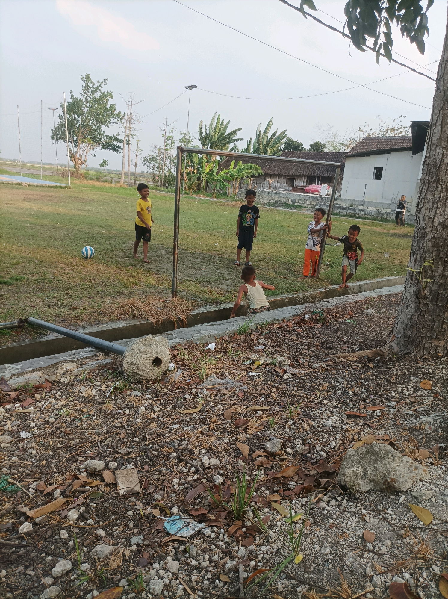 The image depicts an outdoor scene in Sarangan, Indonesia. Several children are present in a grassy area with a soccer goal. A young boy in a yellow shirt and black shorts is standing to the left of the goal, looking forward. Another boy in a black t-shirt with a graphic print is to the left of the goal as well. To the right of the goal, three more children are visible: one in a patterned t-shirt and orange pants, another in a dark t-shirt and patterned shorts, and a third child in a white tank top is partially submerged in a concrete drainage ditch. A soccer ball is on the grass to the left of the goal. The environment includes trees, buildings with tiled roofs in the background, and a large tree trunk on the right side of the frame. The foreground is covered in dry leaves, rocks, and debris. The sky is overcast.