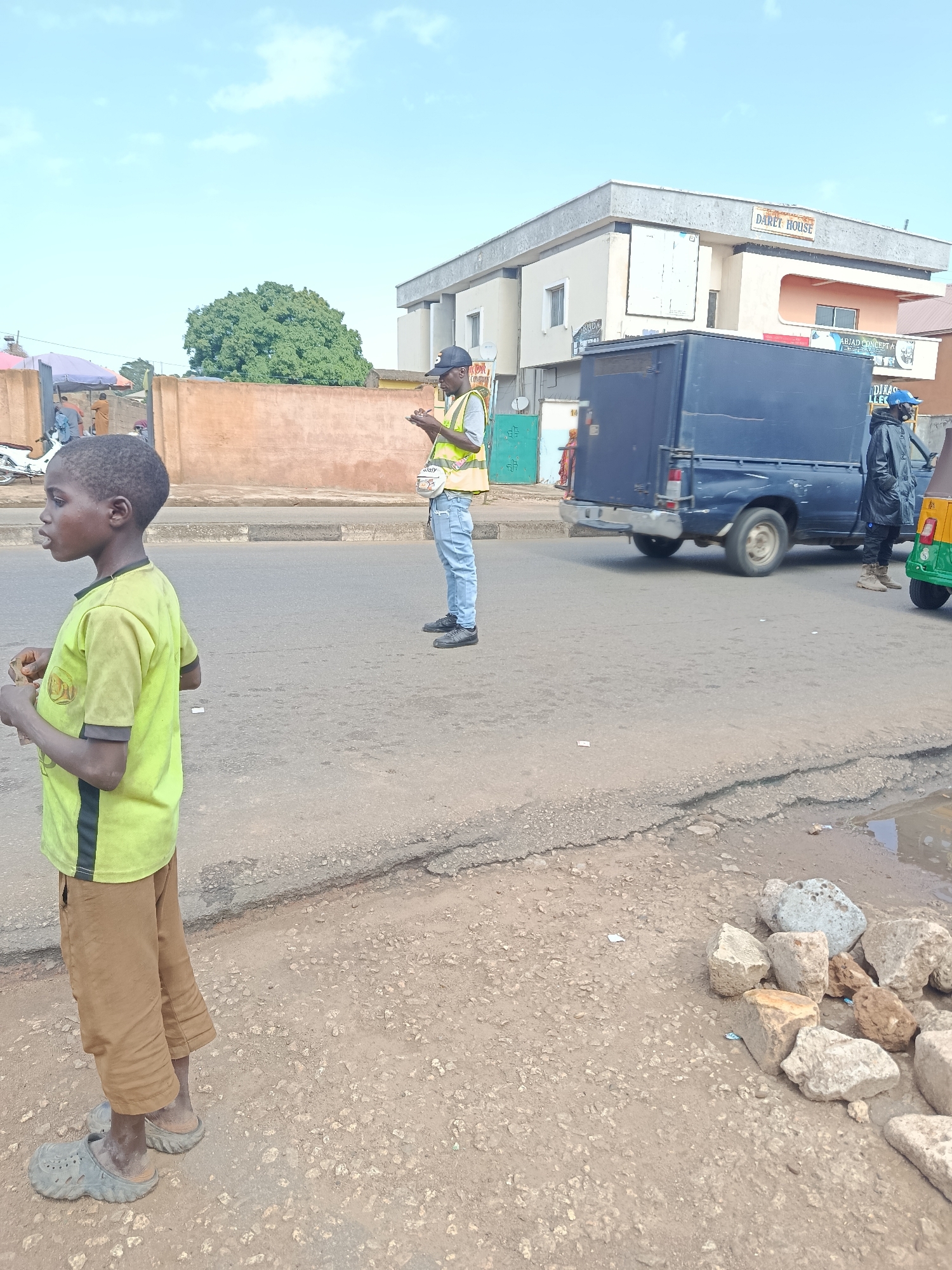 In Jos, Nigeria, on a paved road with significant cracking and pooling water, a young boy in a light green t-shirt and brown pants stands facing left, looking off-camera and holding a small object. In the middle ground, a man wearing a reflective yellow vest and a black cap stands facing the boy, holding his hands up as if in conversation or directing traffic. Behind him, a blue truck and a small, multi-colored vehicle are parked on the side of the road. The background features a tan wall, a large green tree, and a two-story building with "DARET HOUSE" signage. The sky is bright with scattered clouds.