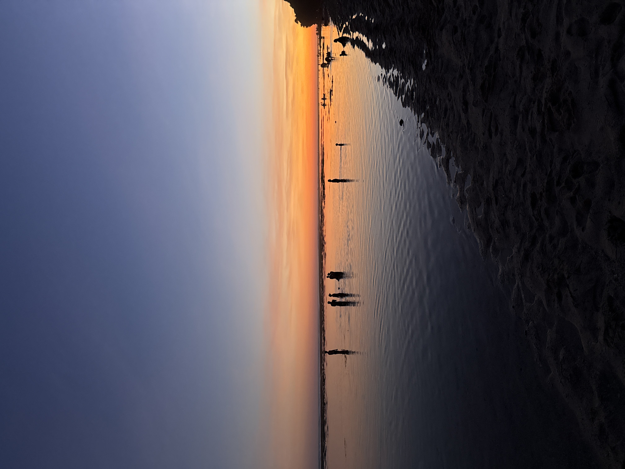 The image captures a sunset over the ocean at a beach in Kuta Selatan, Indonesia. The sky transitions from dark blue to orange at the horizon. The water reflects the colors of the sky. Several silhouettes of people are visible wading in the water. Dark, textured sand borders the water's edge. The image is rotated 90 degrees counter-clockwise.