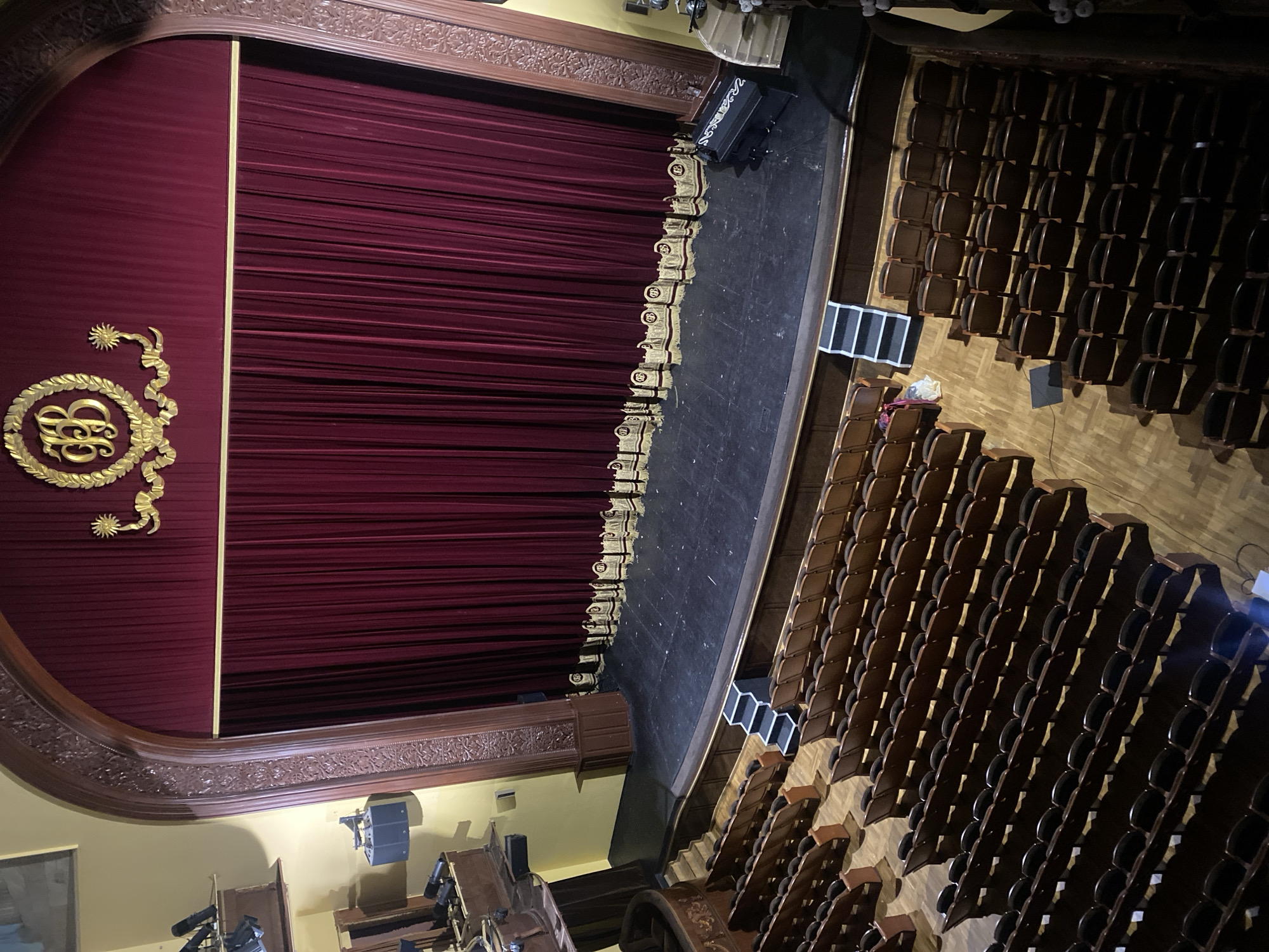 The image depicts the interior of a theater in Заволжье, Russia. A red curtain is closed on the stage, adorned with gold-colored ornamental designs. The stage is black. Rows of wooden seats fill the auditorium, which appears mostly empty. The theater's interior is lit by artificial lighting.