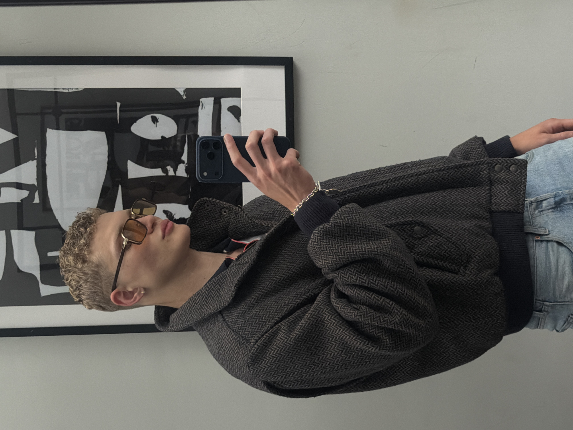 A young person with short, blonde, curly hair and sunglasses is taking a selfie in a room. They are wearing a dark herringbone pattern bomber jacket over a dark shirt, and light blue jeans. A silver bracelet is visible on their left wrist. The background shows a large framed black and white abstract artwork on a light-colored wall. The lighting suggests an indoor environment, possibly a dressing room or hallway. There is no discernible information to indicate the location is in London, United Kingdom.