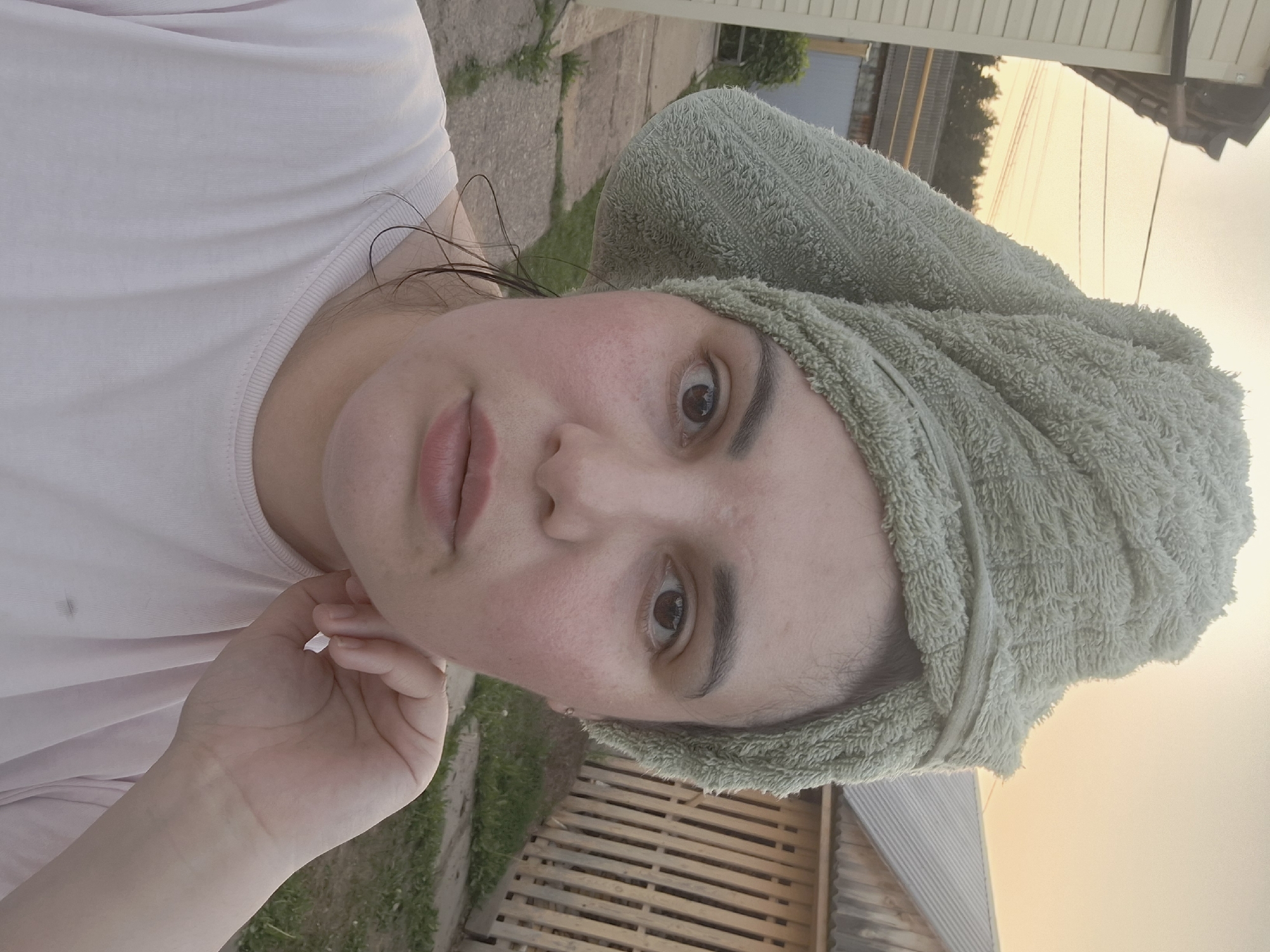 A person with fair skin is depicted in a selfie-style photograph with a green towel wrapped around their head. They are wearing a light-colored shirt and holding their hand near their face. The background includes vegetation, a wooden structure, and a house. The image was captured outdoors, likely in the city of Амзя, Russia.