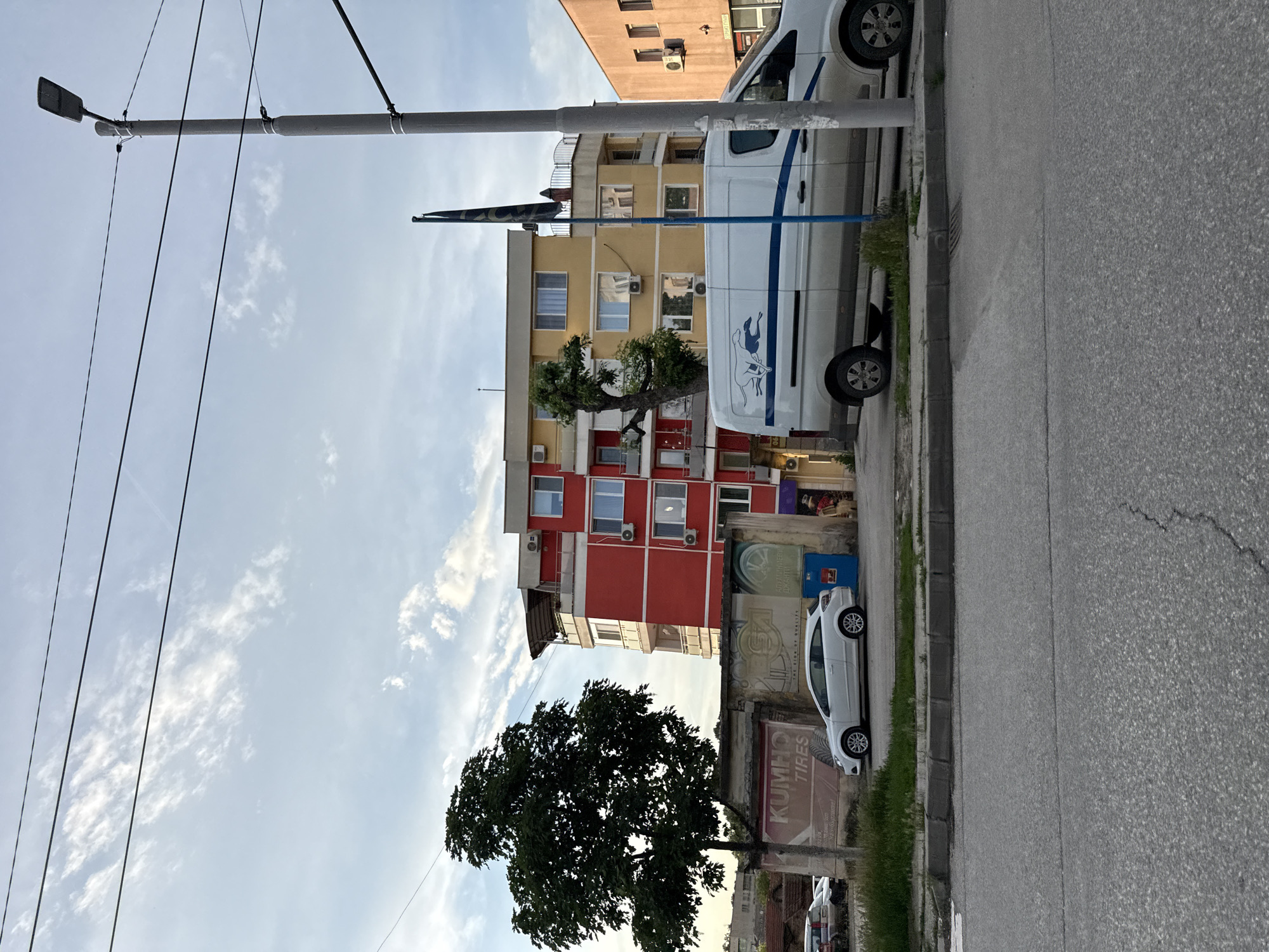 The image depicts a street scene in Pleven, Bulgaria. A utility pole with overhead wires is visible on the left, extending into the sky with scattered clouds. Buildings are situated to the right of the pole, including a structure with a red lower section and a yellow upper section. A tree partially obscures the front of the building.
A white van is parked along the curb, obscuring a portion of the building. Further down the street, a white compact car is parked next to a sign that reads "KUMHO TIRES." The road stretches into the distance.
