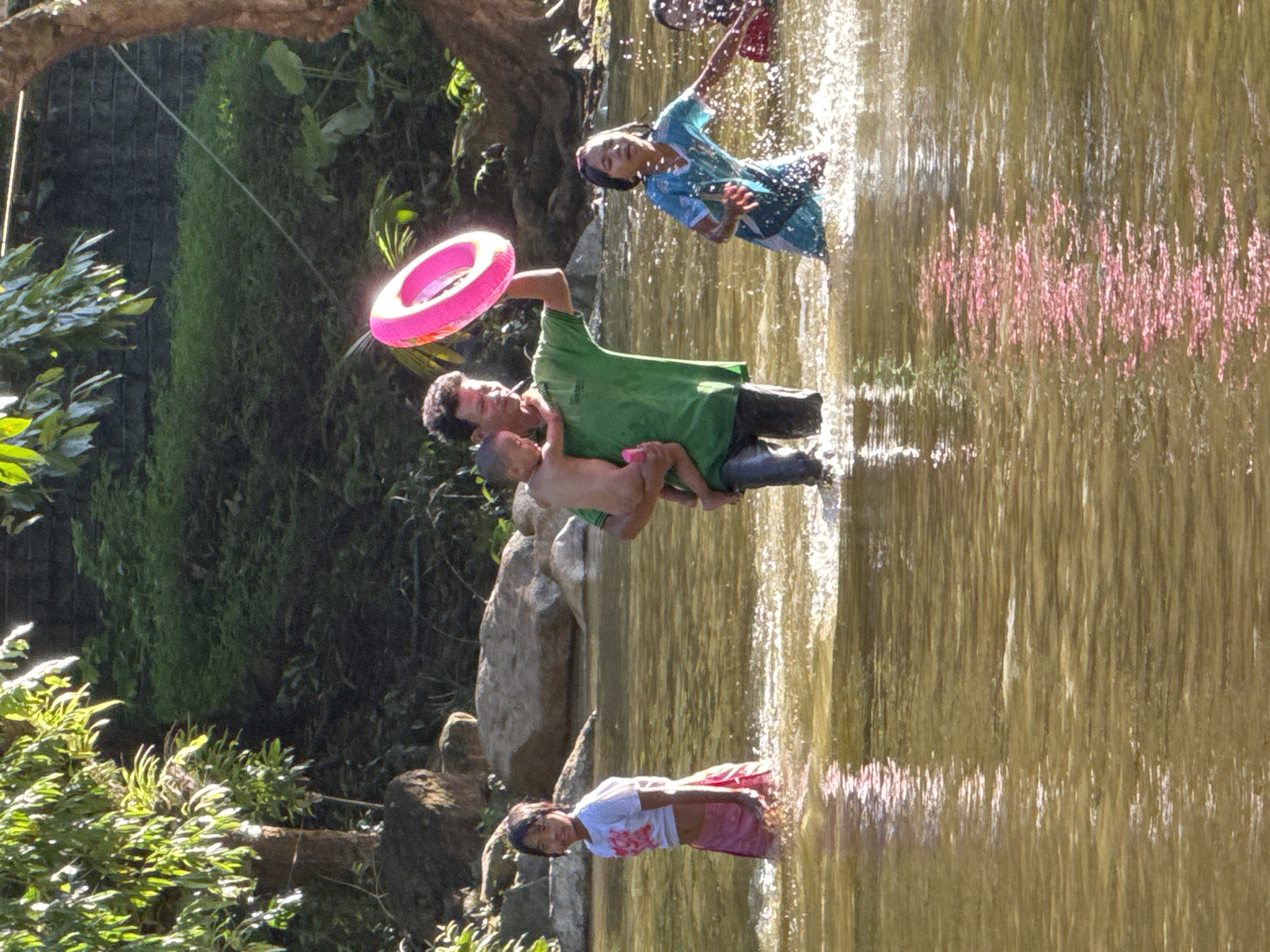 In Ranong, Thailand, a group of children and a man are wading in a shallow, muddy river. Sunlight filters through the dense foliage lining the riverbank, creating dappled patterns on the water's surface. A man in a green shirt carries a young child in his arms and holds a pink inflatable ring aloft. To his left, a young girl in a white shirt and pink skirt stands facing the camera. To the right, a girl in a blue and green patterned dress splashes in the water, with another child partially visible in the background. The riverbed appears to be composed of sand and rocks. The overall impression is one of a casual, recreational outing in a natural, tropical setting.