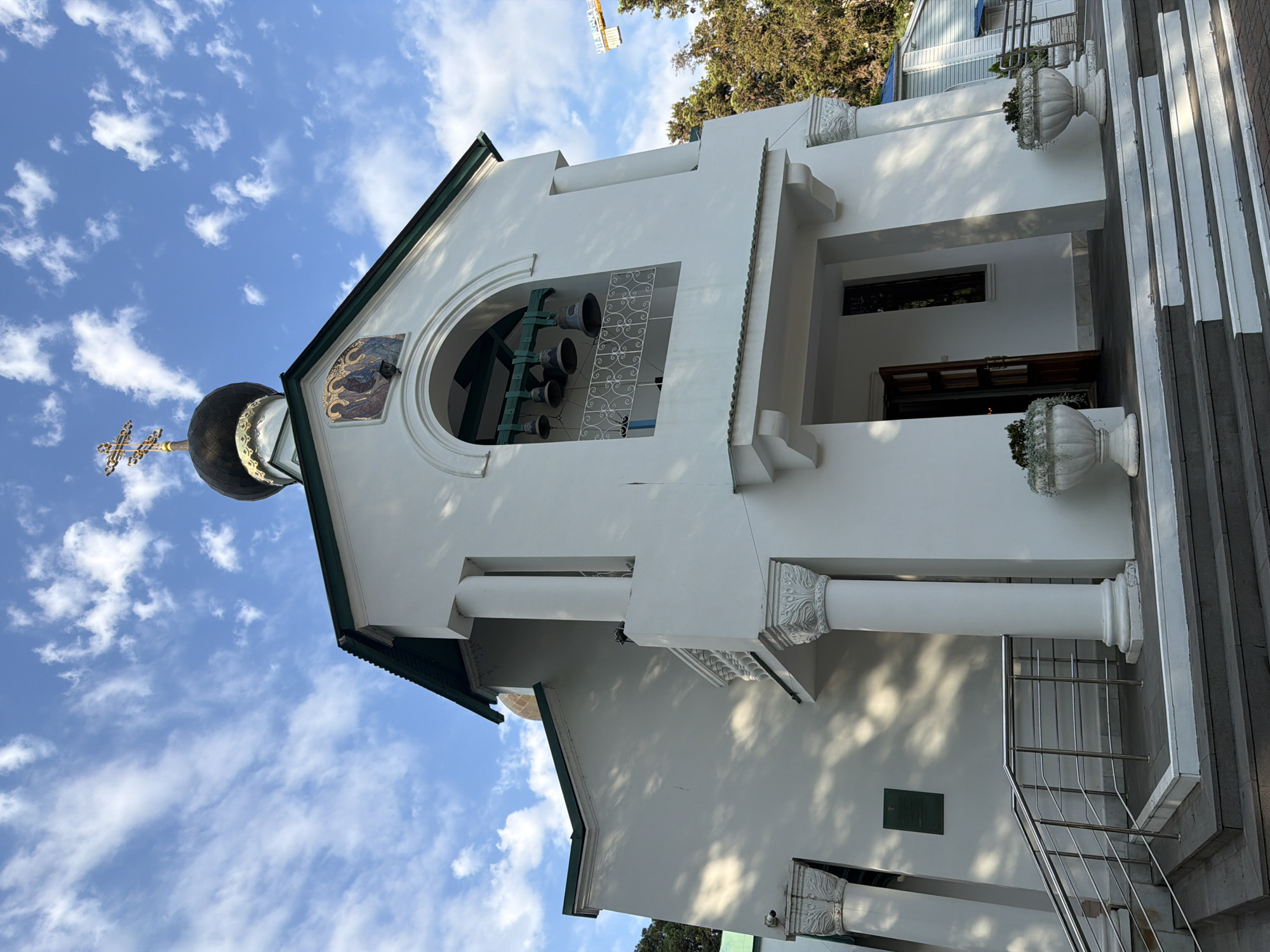 The image shows the bell tower of an Orthodox church in Sochi, Russia, under a partly cloudy sky. The church is painted white with a green roof and a black dome topped with a gold cross. The bell tower has an arched opening with several bells hanging inside. There are decorative columns on either side of the entrance. Two white planters with greenery are situated on either side of the steps leading up to the entrance. A building and trees are visible in the background.