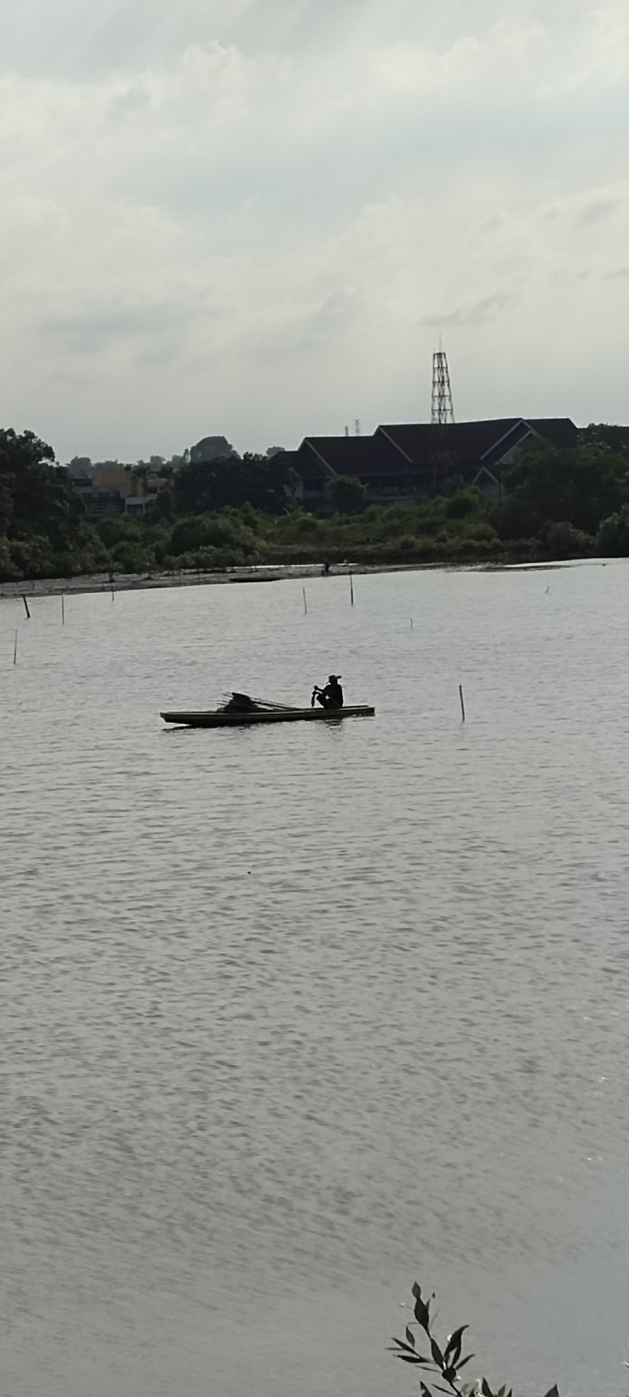 A wide shot captures a serene body of water with a person in a small, flat-bottomed boat in the middle ground. The boat appears to be constructed of wood and is laden with what looks like bundles of reeds or bamboo. The individual is seated in the boat, silhouetted against the lighter background. The water exhibits a subtle rippling texture, suggesting a gentle breeze.
In the background, a distant shoreline is densely lined with green foliage, indicating a natural environment. Behind the trees, a long, dark-roofed building complex is visible, with a tall, lattice-like tower structure situated to its right, reaching into the overcast sky. The sky itself is a soft, diffused grey, characteristic of a cloudy day.
The scene is located near the coast of Lhokseumawe, Indonesia, likely in an area with mangrove forests or aquatic vegetation, given the presence of the water and the dense greenery along the shore. The water body could be a bay, a river estuary, or a large tidal pond within the city's coastal landscape. The overall atmosphere is calm and tranquil.
