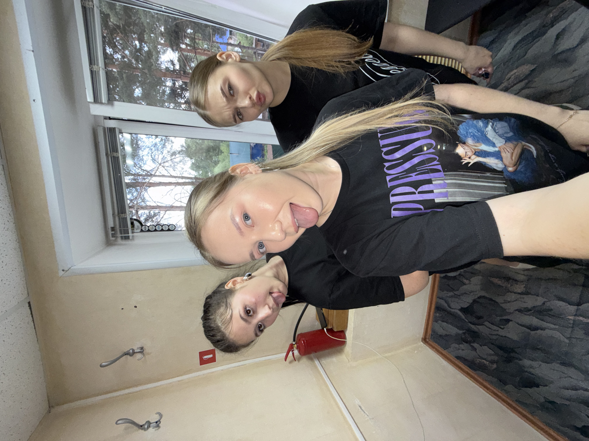 Three young women are posing for a photo indoors. The woman closest to the camera has blond hair and is sticking her tongue out. Behind her and slightly to the left is another woman with dark hair who is also sticking her tongue out. The third woman, positioned higher in the frame, has reddish-blond hair pulled back in a ponytail and is puckering her lips. All three are wearing black T-shirts. In the background, there is a window showing trees outside. A red fire extinguisher is mounted on the wall. Silver coat hooks are also visible on the wall. The overall impression is a playful selfie taken inside a room. The location is not discernable from this photo to be in Chaikovsky, Russia.