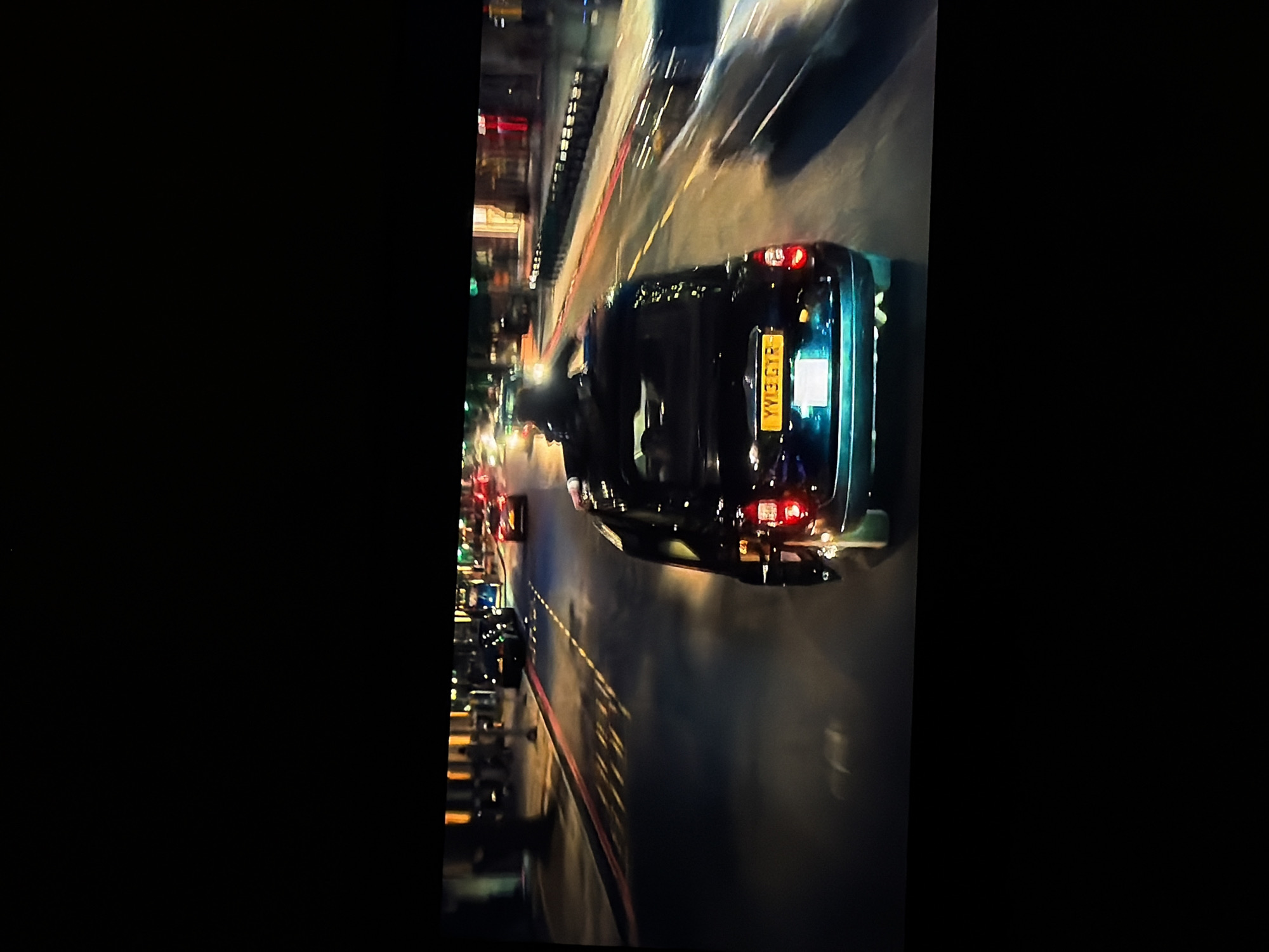 The image, rotated 90 degrees clockwise, depicts a night-time street scene with significant motion blur, suggesting movement. A dark-colored car, appearing to be an SUV or hatchback, is the central subject, viewed from its rear. Its brake lights are illuminated, and faint reflections of surrounding lights are visible on its dark bodywork. A blurry figure, likely a person, is visible near the driver's side of the car, appearing to be interacting with the vehicle, possibly entering or exiting.
The car's rear license plate is clearly visible, bright yellow with black characters, displaying the text "YY13 GYR". This license plate style is characteristic of vehicles registered in the United Kingdom, which conflicts with the provided location context of Sofiivska Borshchahivka, Ukraine.
The road surface beneath the car shows blurred yellow and red markings, indicative of lane dividers or curbs. In the background, numerous streaks of light from streetlights, buildings, and other vehicles create a dynamic urban or suburban atmosphere. The overall impression is one of a busy street at night, captured mid-action. No specific weather conditions are discernible.