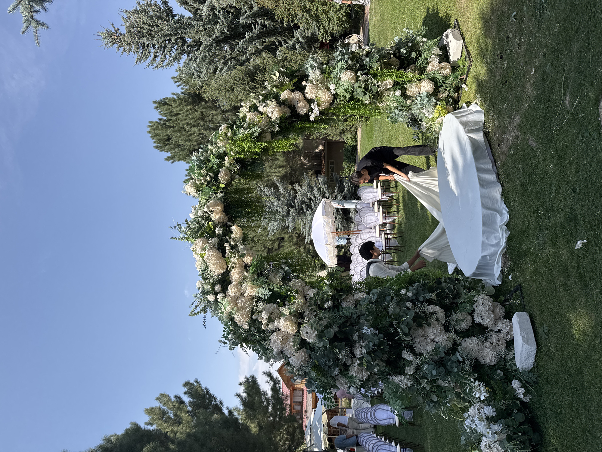 In Koy Tash, Kyrgyzstan, a wedding arch decorated with white flowers and greenery stands outdoors on a grassy lawn. Two individuals are actively arranging white fabric around a circular base beneath the arch. In the background, rows of white chairs are set up, covered with protective white fabric, indicating preparations for an event. Several trees, including pine trees, frame the scene, and a building with a red roof is visible in the distance. The sky is clear and blue.