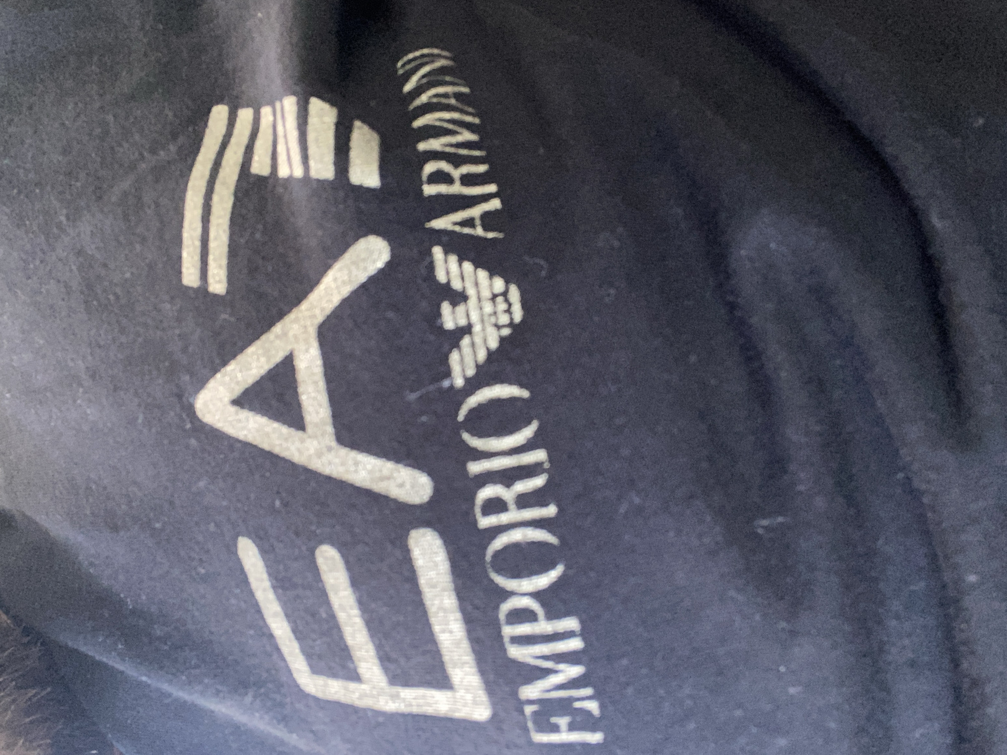 The image shows a close-up of a navy blue fabric, possibly a t-shirt, with a white printed logo. The logo consists of the letters "EAT" in a stylized font, followed by the Emporio Armani eagle emblem and the word "ARMANI". The fabric appears to be cotton and has a slightly textured surface. The image is taken from an angle, with the top left corner showing a hint of dark, possibly fur, material, suggesting it might be part of a garment worn by someone. The background is dark and out of focus, providing no discernible environmental details. There is no information in the image that suggests a location in Varna, Bulgaria.
