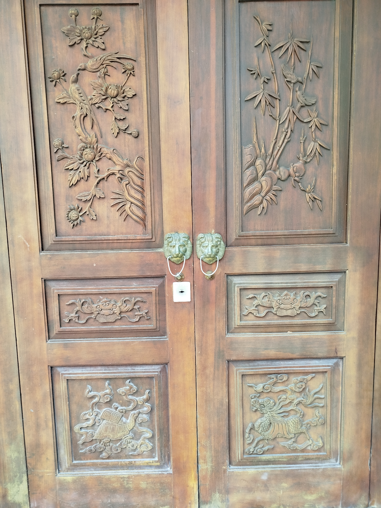 The image shows a pair of ornately carved wooden doors. Each door is divided into four rectangular panels. The upper panel on the left door features a carving of birds amidst flowers and branches. The upper panel on the right door displays a carving of bamboo stalks with birds. The middle panels of both doors have similar carvings of a decorative, stylized design. The lower panels are different: the left door depicts a turtle-like creature amidst swirling patterns, while the right door shows a horse-like creature in a similar swirling environment. Each door has a lion-head doorknocker positioned in the center, with a white, rectangular security plate just beneath the left door's knocker. The doors appear old and weathered, and are located in Ho Chi Minh City, Vietnam.