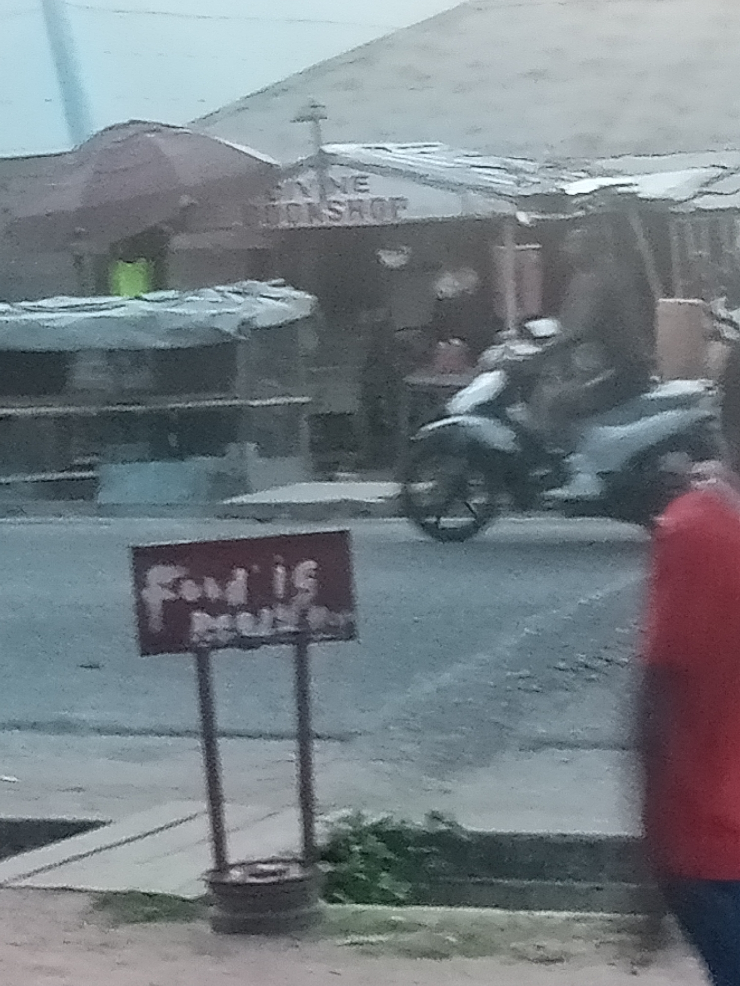 The image depicts an outdoor scene with a building in the background, possibly a workshop or shop, with signage that appears to read "MINE BOOKSHOP." In the foreground, a motorcycle is parked or being ridden, with a person on it. There is a red sign on a pole that is difficult to read but appears to have the text "Food is..." on it. A person in a red shirt is visible on the right side of the frame. The environment appears to be a street in Jalingo, Nigeria, with a slightly overcast sky. The image quality is low, with a blurriness that makes precise identification of details challenging.