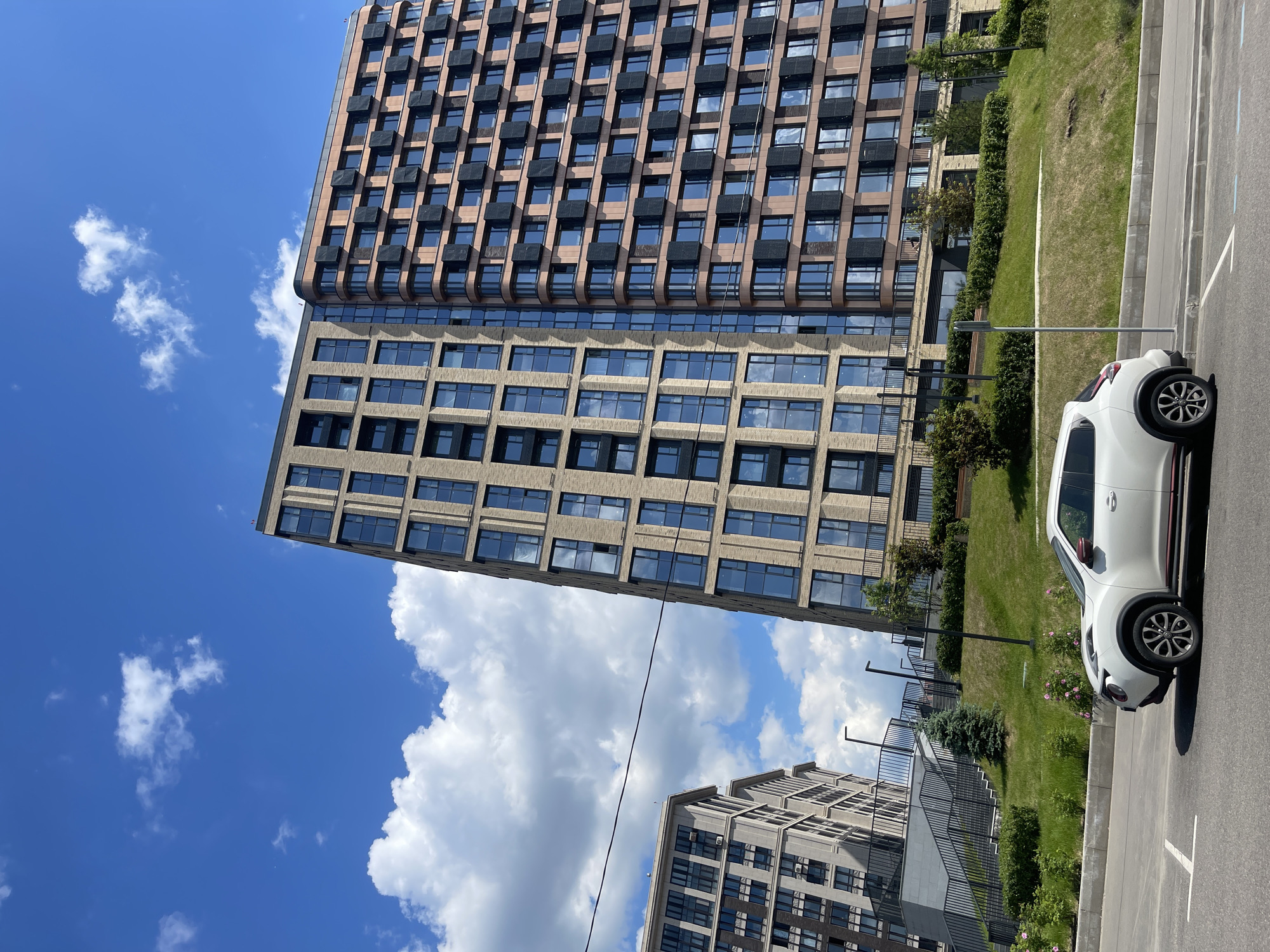 This image captures an urban scene on a clear, sunny day, featuring modern architecture and a parked car.
Dominating the left and central parts of the frame is a tall, contemporary multi-story building. Its upper section is characterized by a facade of brownish-orange panels with rows of dark, projecting elements, possibly small balconies, and recessed windows. Below this, the building transitions to a section with lighter, possibly concrete-colored panels and a grid of large, rectangular windows that reflect the bright blue sky and white clouds. In the background, partially visible on the lower right, is another modern building with a lighter, grayish facade and numerous windows.
Between the buildings and the foreground, a strip of green grass is visible, interspersed with bushes and small trees. A black metal fence or railing separates parts of the green space.
In the bottom right, a white car, resembling a crossover SUV like a Nissan Juke, is parked on an asphalt surface. The car faces towards the right, showcasing its front end, distinctive black wheel arches, and multi-spoke wheels. A white parking line is marked on the asphalt next to the vehicle.
The sky is a vibrant blue with scattered fluffy white cumulus clouds, indicating pleasant weather. Natural sunlight illuminates the entire scene. A thin, dark cable or wire crosses diagonally through the mid-ground. There are no people visible, and no text can be discerned in the image.