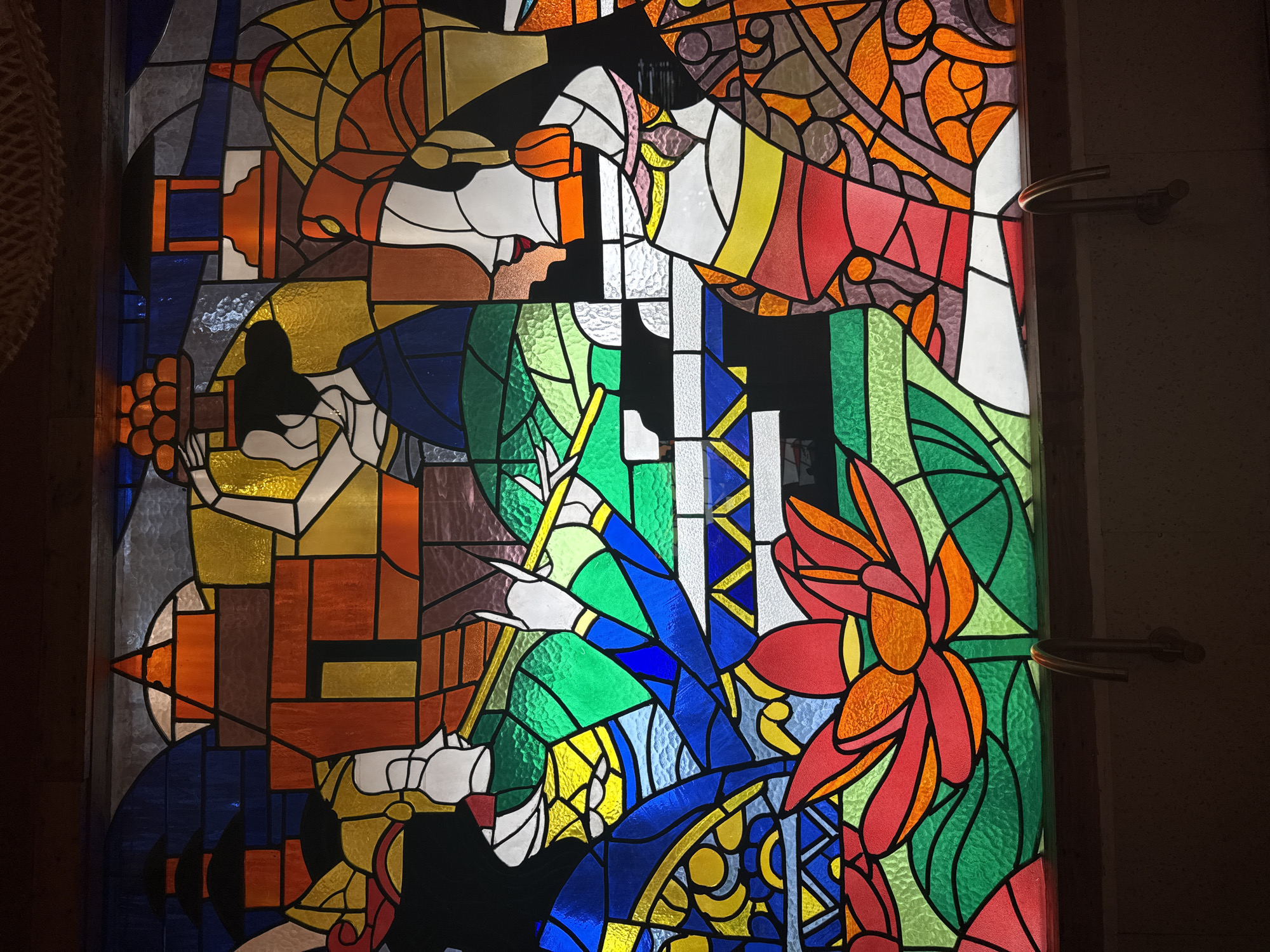 An abstract stained glass window with a multitude of colors including orange, red, green, yellow, blue, brown, and white is depicted. The window contains what appear to be floral designs and geometric shapes. The location is North Kuta, Indonesia.