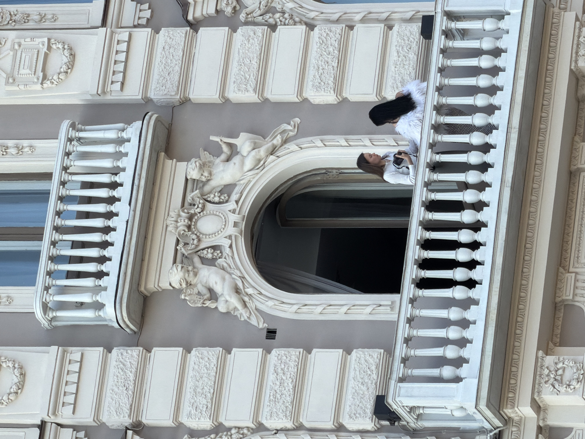 Two women stand on an ornate balcony with a light gray facade. The building is decorated with white trim, including statues of cherubs, balustrades, and patterned rectangular panels. The balcony's opening is arched, with decorative elements around it. One woman, facing the building, is taking a picture, while the other, wearing white feathery clothing, looks out from the balcony. It is likely a building in Saint Petersburg, Russia, judging from the architecture style.