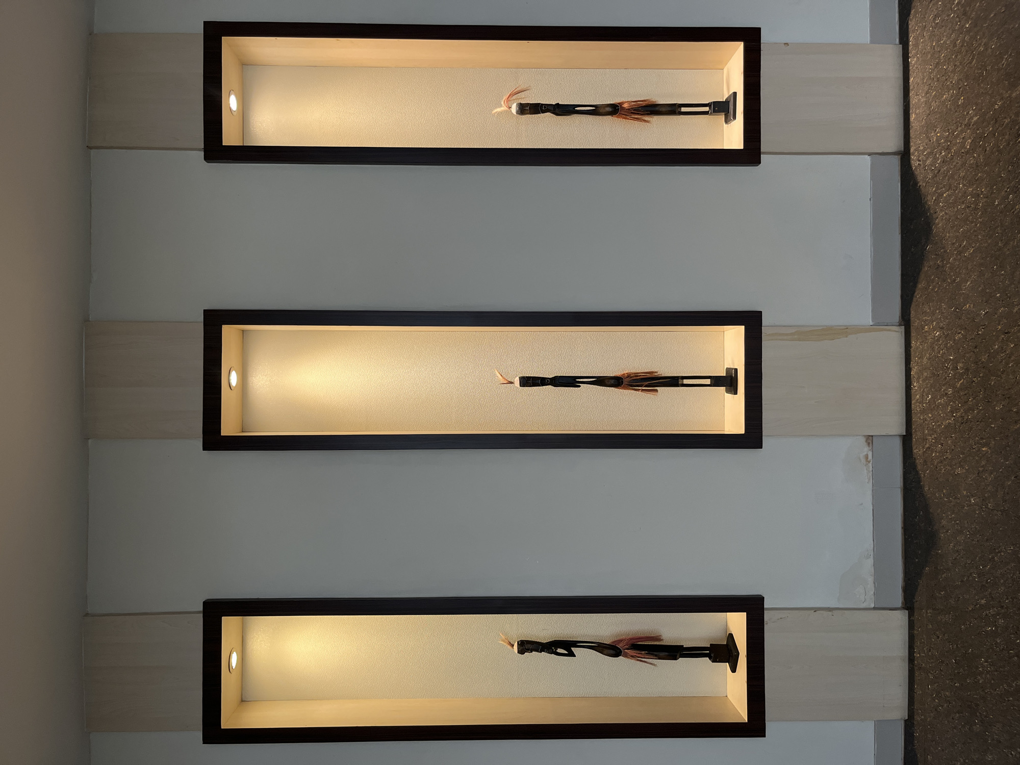 The image displays three recessed display niches on a white wall, each housing a dark wooden figurine. The figurines are slender and abstract, with prominent headdresses made of light-colored feathers and decorative elements around the waist that appear to be made of reddish-brown strands. Each niche is illuminated by a spotlight from above, casting a warm glow. The wall is painted white and has textured panels on either side of each niche that are lighter in color and appear to be made of wood or a similar material. The floor is dark and textured, resembling speckled linoleum or tile. The overall setting suggests an indoor display area, possibly a lobby or hallway. The location is identified as Kuta Selatan, Indonesia.