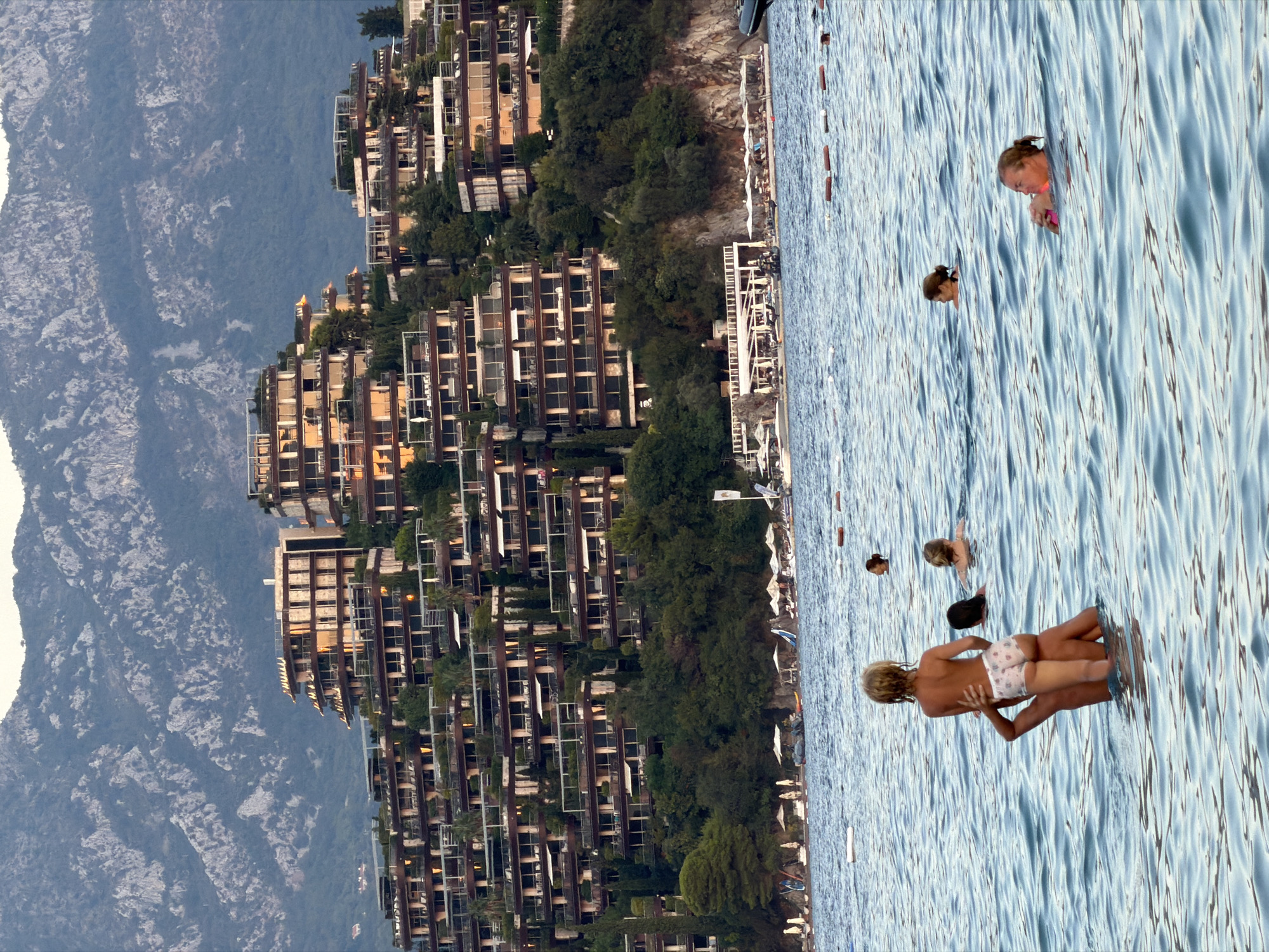 In the foreground of the image, several people are swimming in the sea. A man is carrying a young child on his shoulders, the child facing away from the viewer. To the right, a woman is swimming and holding onto a pink inflatable object. Further back, another woman is swimming. The sea is blue with gentle waves.
In the midground, a beach area is visible with numerous white umbrellas and lounge chairs. Several boats are docked along the shore, and a large, modern resort complex is built into the hillside behind the beach. The resort features multiple levels with balconies and dark-framed windows, appearing to cascade down the slope.
The background is dominated by a large, rocky mountain range with patches of vegetation. The sky is a soft, muted color, suggesting it is either early morning or late evening. The overall scene depicts a resort area in a coastal mountainous region, likely Budva, Montenegro, known for its attractive beaches and hillside developments.