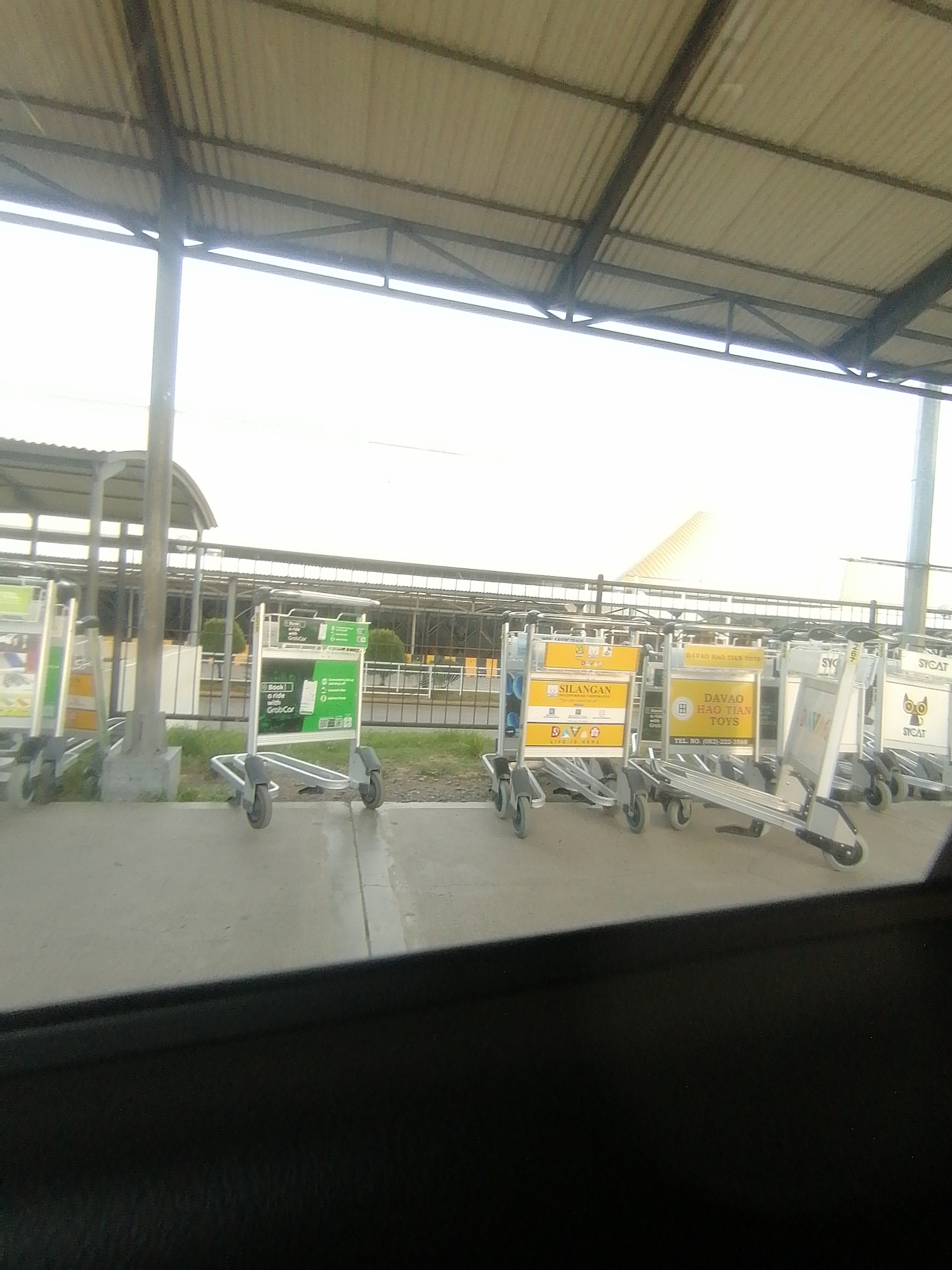 The image depicts an outdoor area at what appears to be an airport, likely in Davao City, Philippines, as indicated by visible signage. The main subjects are numerous silver airport baggage carts or trolleys, neatly arranged on a concrete surface in the foreground and mid-ground.
Several carts feature advertising panels. One prominent panel displays "Book a ride with GrabCar" in green. Other carts show "SILANGAN" and "DAVAO HAO TIAN TOYS" with a telephone number "(082)-222-3568," directly referencing Davao. Partially visible text such as "SYCAT" and "DVAO" also appears on some carts.
Above the carts, a large, corrugated metal roof structure, supported by metal beams and pillars, extends across the upper portion of the frame, suggesting a covered area for passengers or a sheltered walkway. Beyond the carts and a metal fence, a large building with a distinctive peaked roof is visible against a bright, washed-out sky, strongly resembling an airport terminal.
The scene suggests an environment associated with travel and transportation. No people are visible, but the presence and arrangement of the baggage carts imply an expectation of passenger activity, such as arrivals or departures. The lighting indicates daytime, possibly an overcast sky or bright conditions causing some overexposure. The ground beneath the carts is concrete, with patches of green grass visible in the background. The overall impression is one of an organized, functional airport facility.