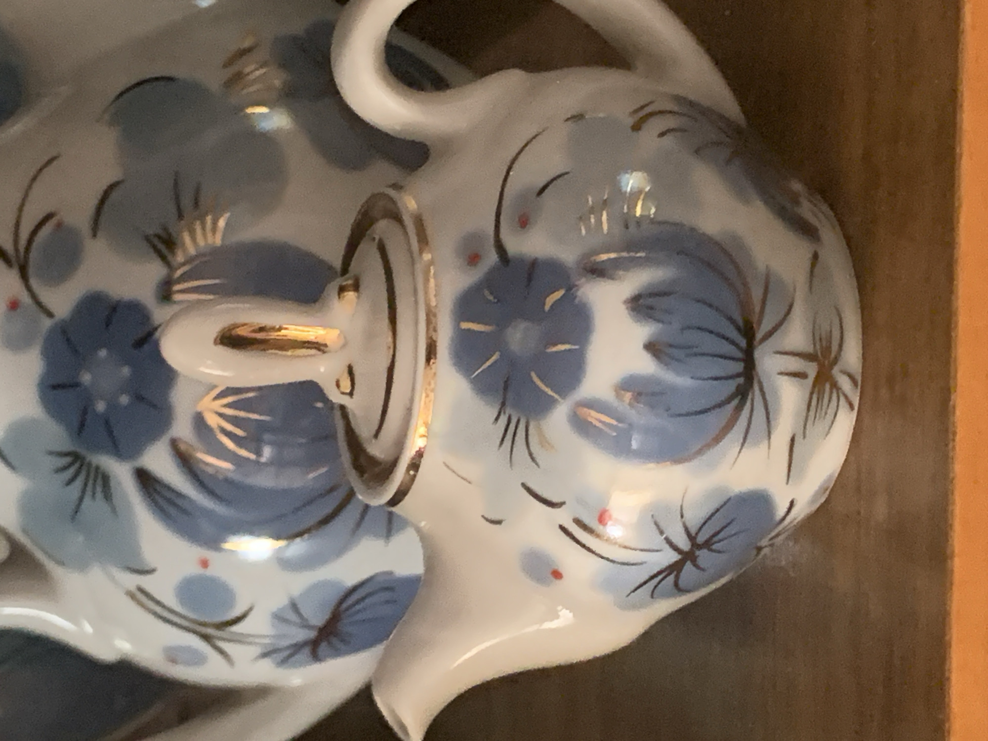 A close-up, eye-level, indoor shot features a small, decorative white teapot adorned with blue floral patterns and gold accents. The teapot is positioned on a dark wooden surface. In the background, a larger, similarly decorated teapot is partially visible. The lighting is warm and highlights the reflective surfaces of the porcelain and gold trim. The image does not contain any people or provide any information about its location in Tashkent, Uzbekistan, or any other city.